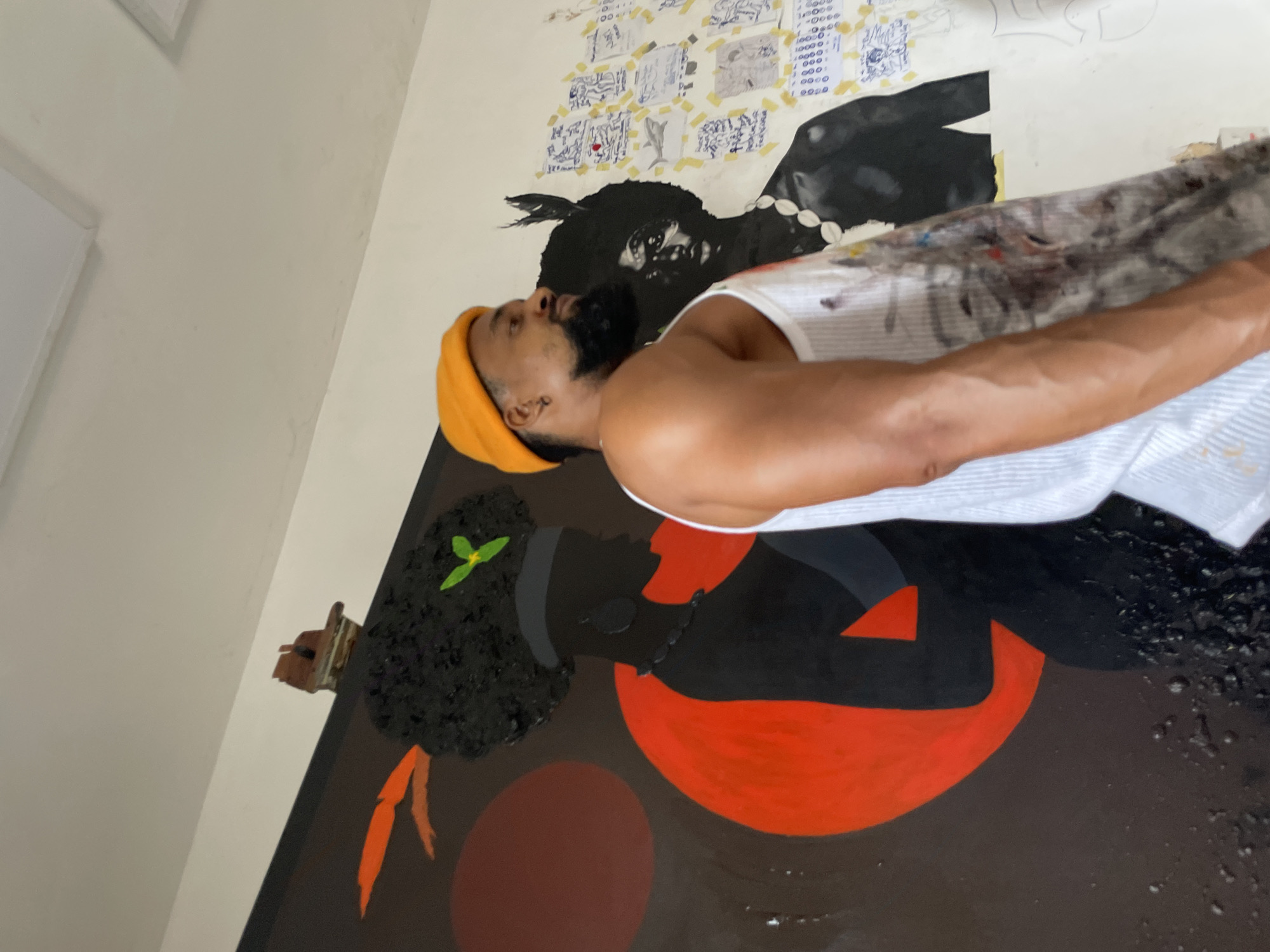 A man wearing a yellow beanie and a white tank top stands in front of a large painting. The painting depicts a black man with a beaded necklace. Above the figure in the painting, there are several papers taped to the wall with drawings and writing on them. The location is possibly Ado, Nigeria. The man appears to be an artist working on the painting. The painting itself is composed of black, red, and brown colors with the image of a kneeling person in the lower right and a black figure in the upper right.