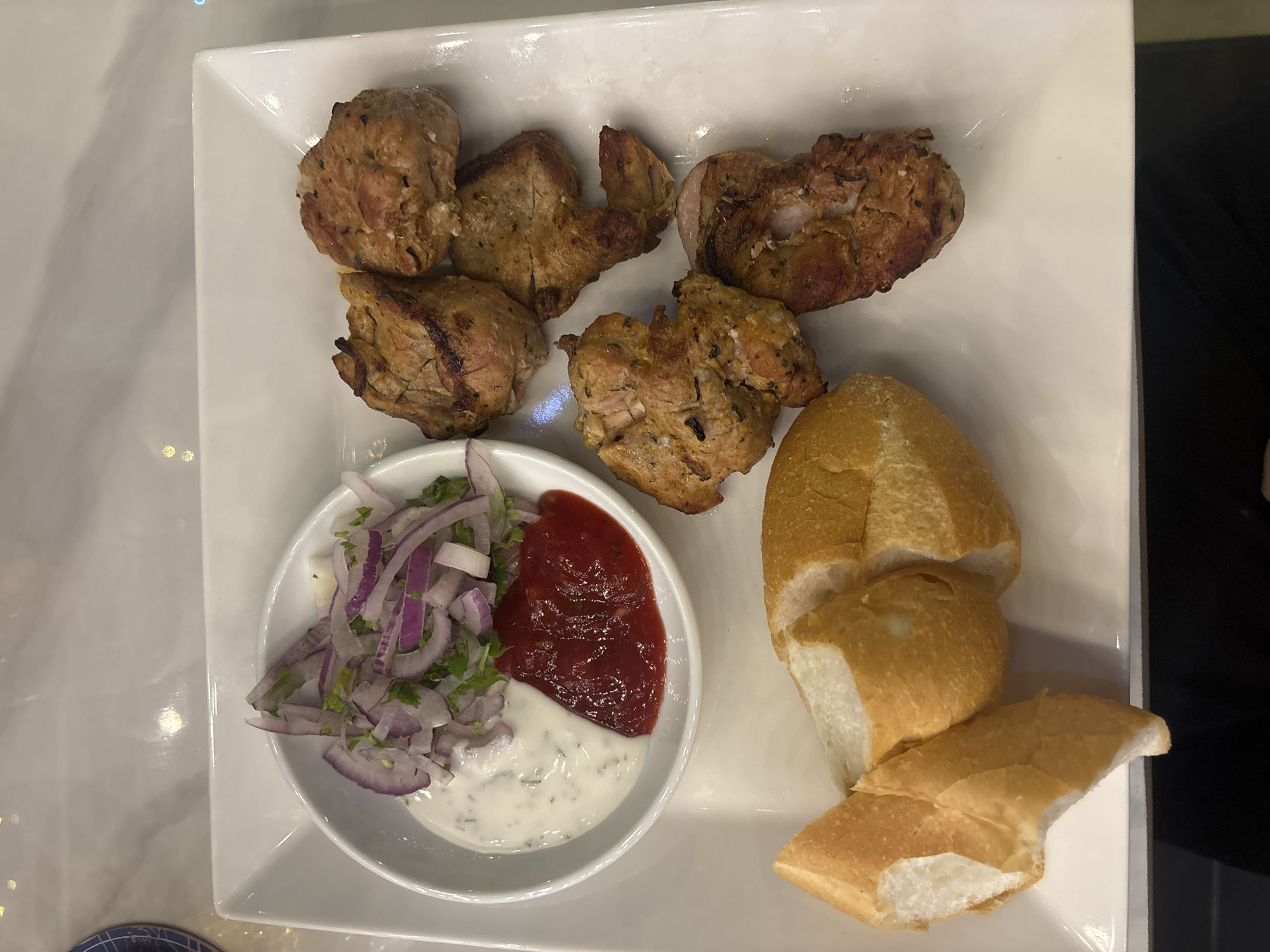 A close-up, overhead shot captures a white, square plate of food. The plate contains grilled meat skewers, a small white bowl filled with a creamy white sauce and red sauce, sliced bread, and a garnish of chopped red onion and parsley. The meat skewers are arranged on the right side of the plate, with six pieces visible. The white bowl is located on the upper left side, and the sliced bread is positioned on the lower left. The background appears to be a white marble countertop.