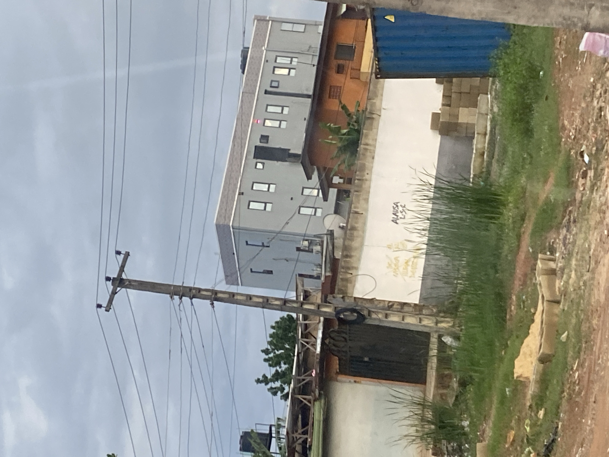 The image depicts a cloudy sky over a street scene in Ikorodu, Nigeria. A utility pole with multiple wires stands prominently in the foreground, leaning slightly to the left. Behind the pole, a multi-story gray building with numerous rectangular windows is visible. In front of the gray building, a smaller, orange-roofed structure is situated. A white wall with some graffiti, including the word "ALAUSA," runs across the mid-ground. Green grass and some debris are scattered on the ground. A large blue shipping container is parked to the right of the wall. The overall impression is of a developing urban area with a mix of residential and commercial structures.