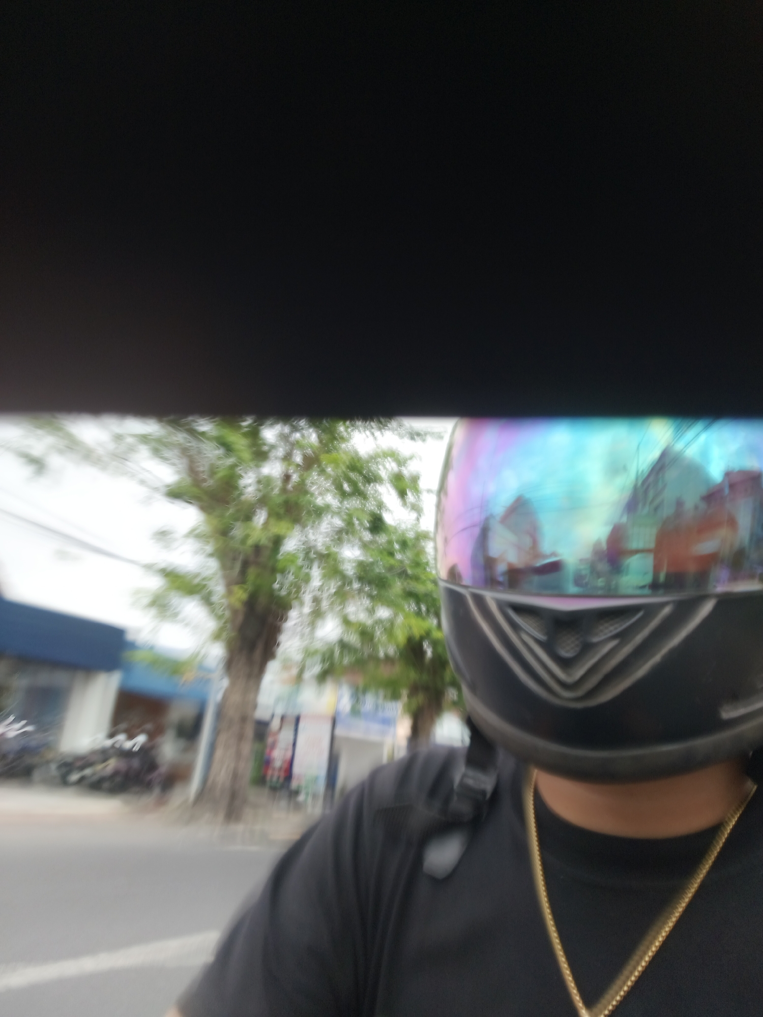The image depicts a first-person perspective of a person wearing a black t-shirt and a motorcycle helmet with a reflective visor. The visor displays a colorful, iridescent reflection of the surroundings. In the background, there is a blurred view of a street scene in Denpasar, Indonesia. Visible elements include a tree with green foliage, a building with a blue facade, parked motorcycles, and street signs. The motion blur suggests the person is likely riding a motorcycle or in a moving vehicle. The overall impression is a candid, in-the-moment snapshot of travel in an urban environment.