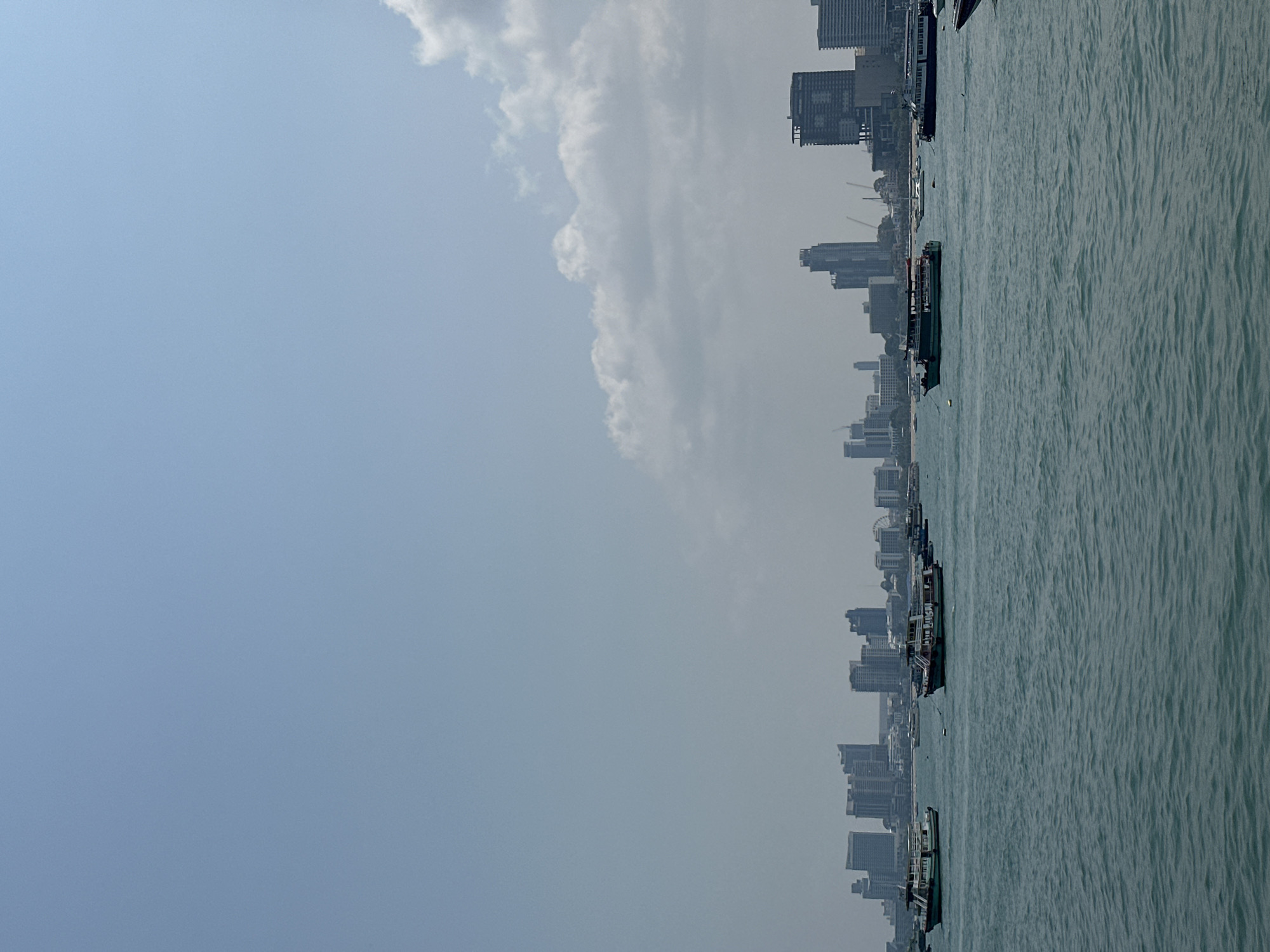 This image depicts the waterfront of Pattaya City, Thailand, under a bright, slightly hazy sky with scattered clouds. The foreground is dominated by the sea, its surface rippled by gentle waves. Numerous boats, possibly fishing vessels or tourist boats, are anchored or moored close to the shore. Along the shoreline, a dense cityscape stretches into the distance, comprising buildings of varying heights, including high-rise condominiums and hotels. Construction cranes dot the skyline, suggesting ongoing development. The overall impression is a bustling urban coastal environment.