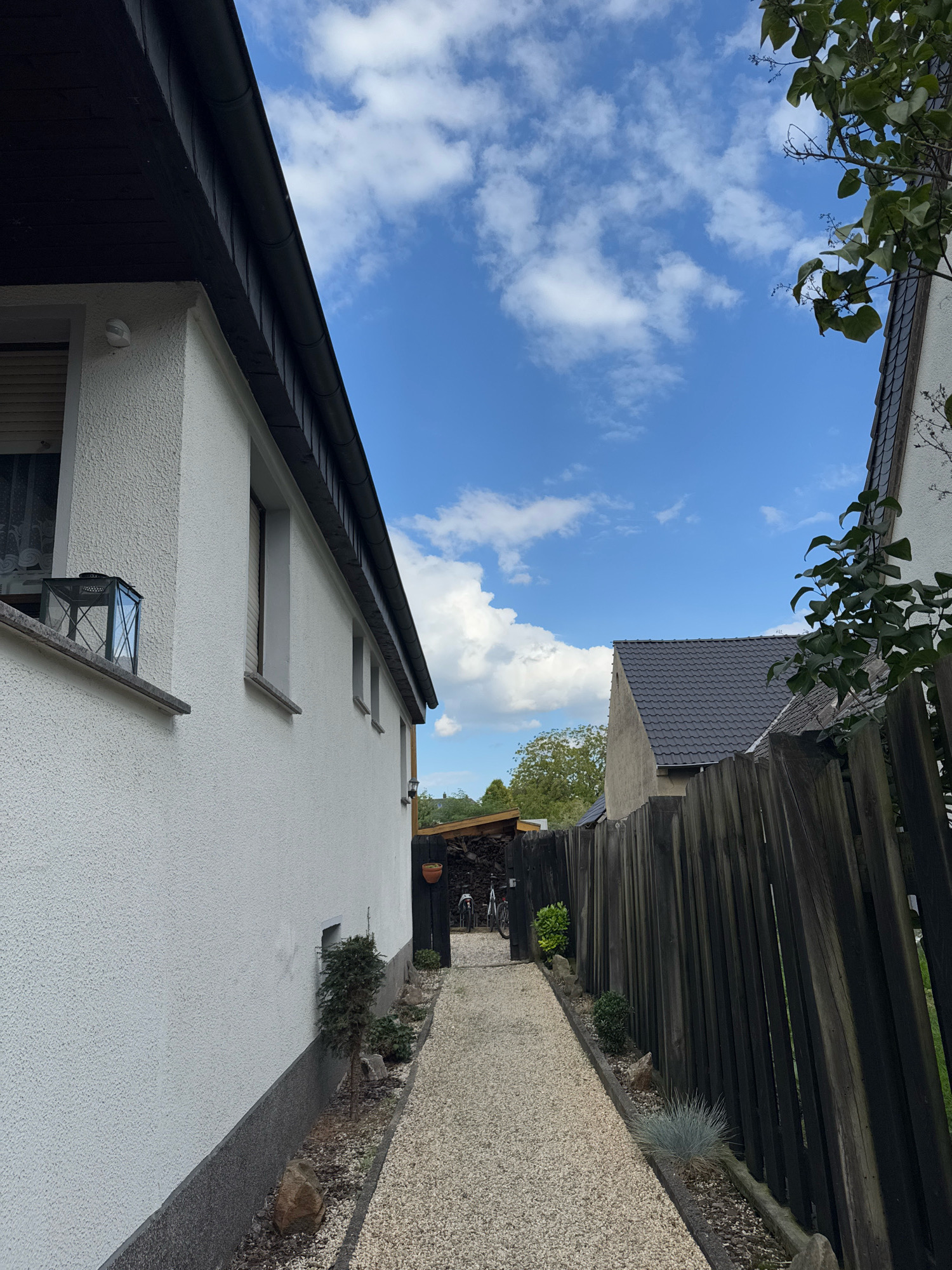 The image depicts a narrow gravel pathway leading between two houses, with a wooden fence on the right. The pathway is bordered by small plants and rocks. The house on the left is white with a dark roof and features a window with a lantern on its sill. The house on the right has a dark tiled roof and a wooden fence. The sky above is blue with scattered white clouds. In the background, at the end of the pathway, there is a covered area with bicycles. The scene appears to be a residential backyard or alleyway in Cleves, Germany.