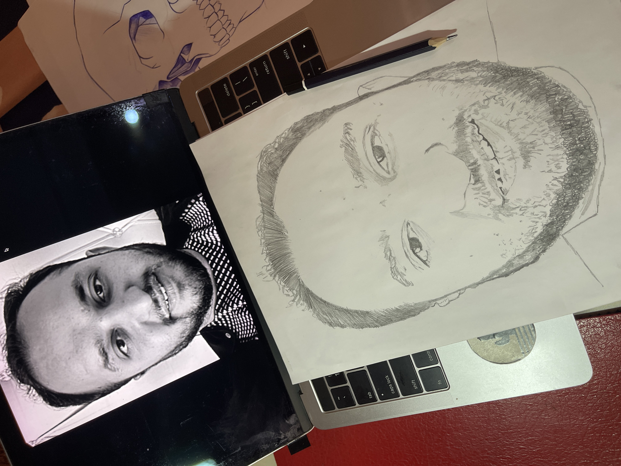 The scene depicts an artist's workspace. A grayscale image of a man's face is displayed on a tablet. A sketch of the same man's face, rendered in graphite, lies on top of a laptop keyboard, with a pencil resting nearby. An additional sketch of a skull is partially visible in the upper left corner. The scene appears to be indoors, set on a red surface. The location is Jos, Nigeria. The image captures a creative process involving portraiture.