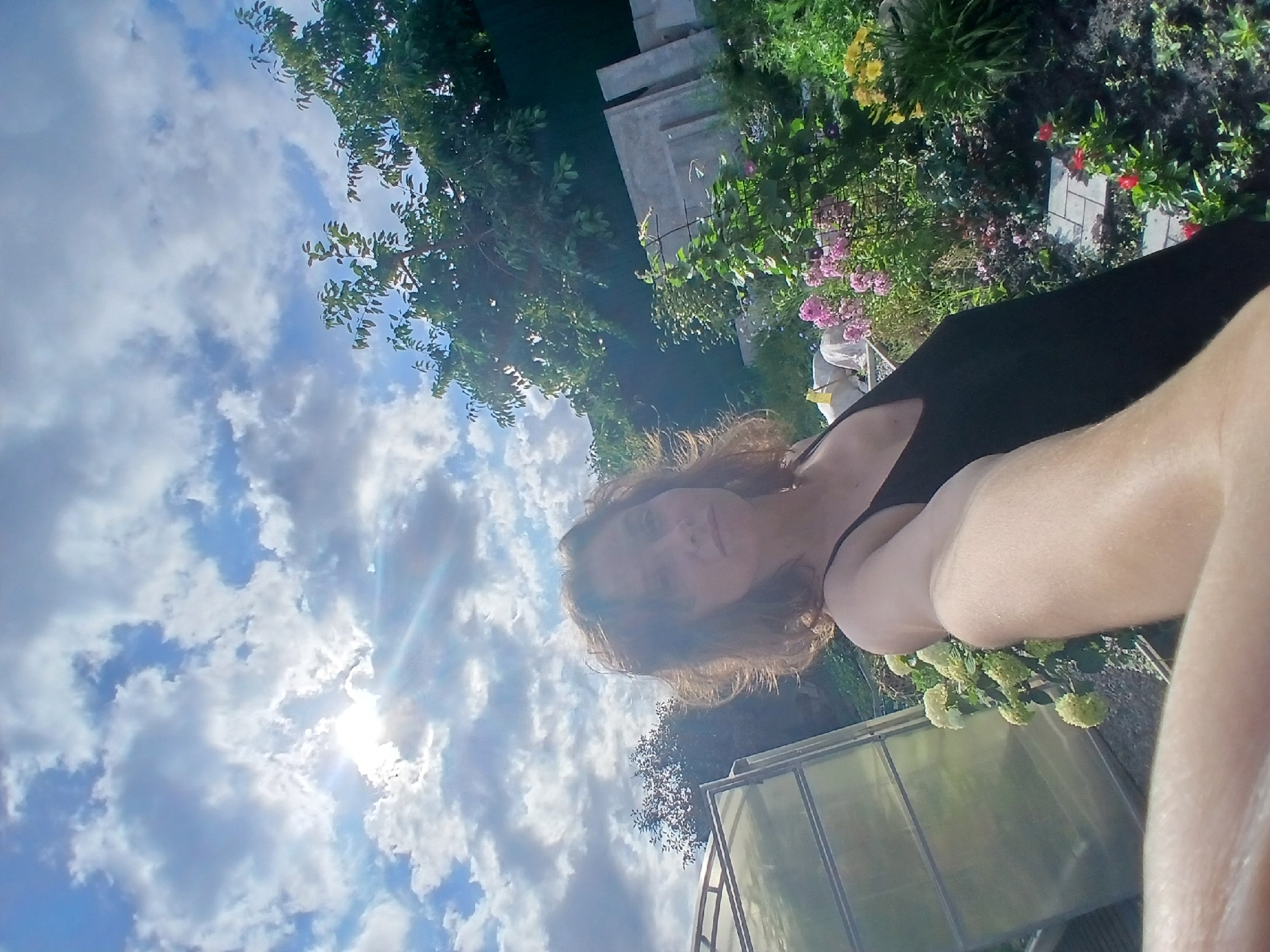 In a candid outdoor shot, a woman with shoulder-length brown hair wearing a black tank top is captured from the chest up, holding the camera in a selfie pose. She is positioned slightly to the right of the frame, her gaze directed towards the viewer. Behind her, the scene unfolds in a garden setting. To the left, a greenhouse with a translucent covering is visible. The garden bed features a variety of blooming flowers in shades of pink, yellow, and red, interspersed with lush green foliage. In the background, trees with dense green leaves and a dark green fence create a natural backdrop. The sky above is a dynamic display of blue with scattered white clouds, and the sun peeks through, casting rays of light. The overall impression is of a warm, sunny day in a cultivated outdoor space. The image may have been taken in the city of Maslova Pristan', Russia, given the context.