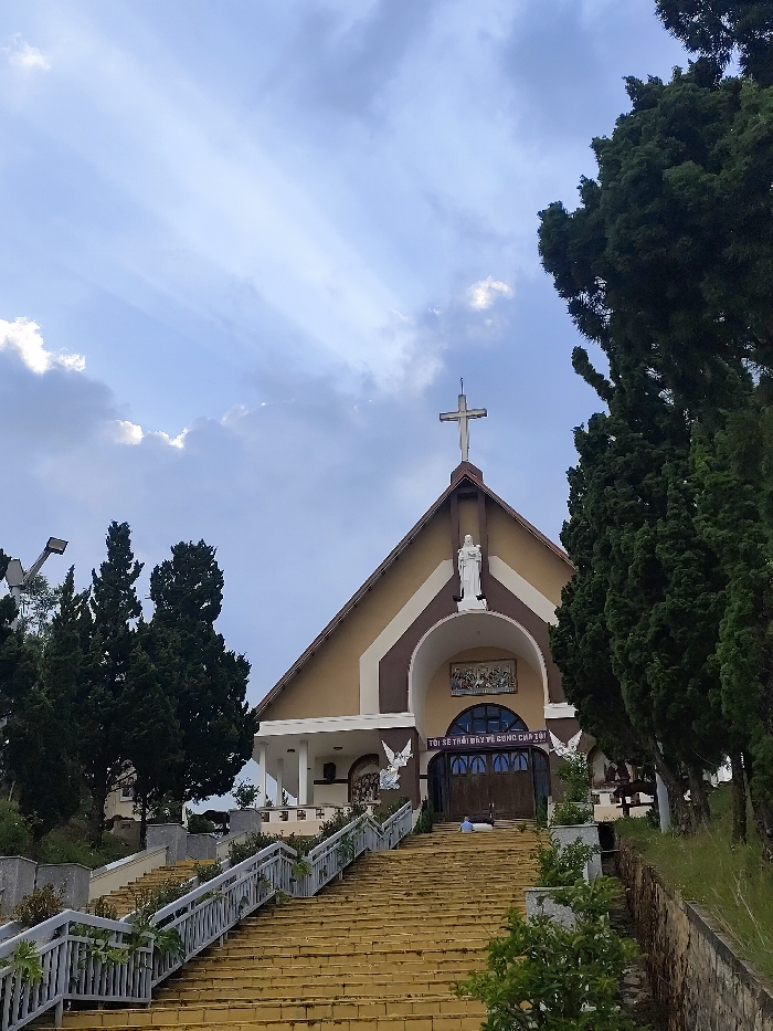 This image depicts the facade of a church, identified as a religious structure within the city of Dalat, Vietnam, due to architectural style and visible Vietnamese text. The church features a steep, pitched roof surmounted by a white cross. The building's exterior is characterized by light brown and dark brown (or reddish-brown) panels, forming geometric patterns around a large central archway.
Above the main entrance arch, a white, robed statue of a figure stands. The arch frames dark double doors, above which a colorful mosaic or mural is visible. A white sign with Vietnamese text, "TÔI SẼ THỜI ĐÀY TẾ CUNG CHẠI" (I will live in the temple), is affixed horizontally above the doors. Two white winged angelic figures are mounted on the facade, flanking the entrance at a lower level. To the left of the main entrance, a smaller, white-pillared portico is visible.
A broad, yellow-tiled staircase ascends directly to the church entrance, flanked by gray metal railings with integrated planters containing green foliage. Along the staircase and the periphery of the scene, numerous tall, dark green coniferous trees (possibly cypress or pine) are present. Patches of green grass and shrubs are visible on either side of the stairs.
One individual, dressed in a light blue top and dark trousers, is situated on the steps directly in front of the church doors, appearing to be sitting or kneeling.
The sky overhead is partly cloudy, with visible shafts of sunlight (crepuscular rays) breaking through clouds in the upper left quadrant of the image. A streetlamp on a tall pole is discernible behind trees on the far left. The scene suggests a tranquil, elevated setting.