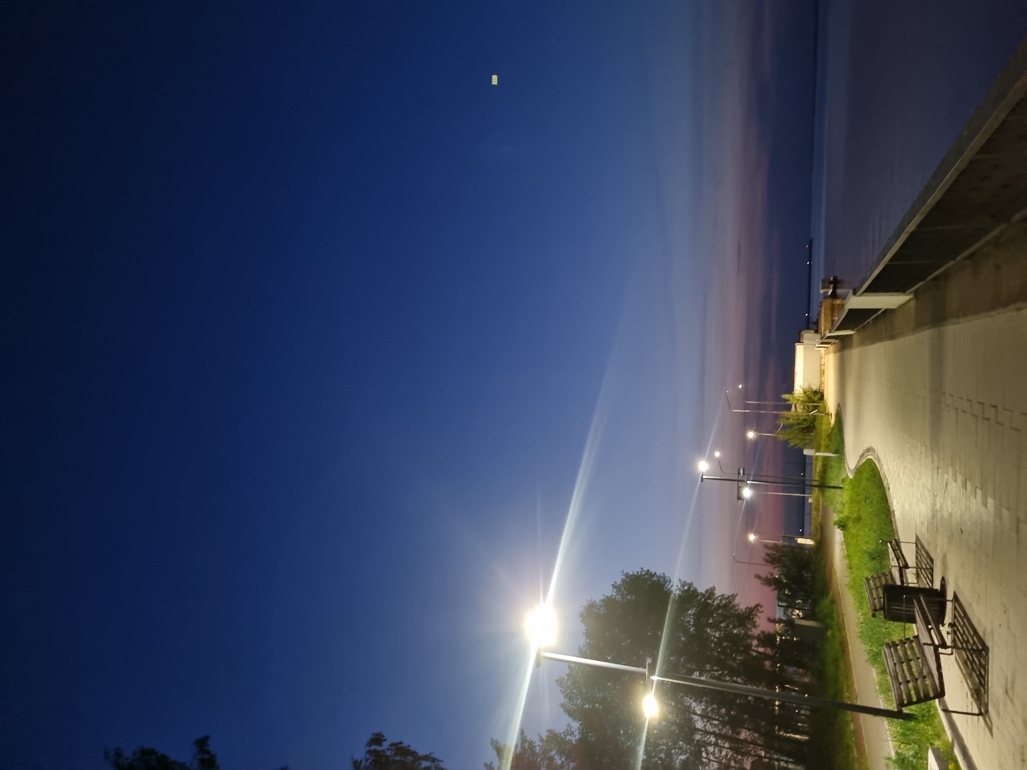 The image captures a nighttime scene on a walkway in Chaikovsky, Russia. The sky transitions from a deep blue to hues of pink and purple near the horizon, where it meets a body of water. Streetlights illuminate the walkway, casting a bright glow and creating long shadows. Benches line the path, suggesting a place for relaxation. Trees and other vegetation are visible along the walkway. A concrete barrier runs along the edge of the water, providing a sense of enclosure.