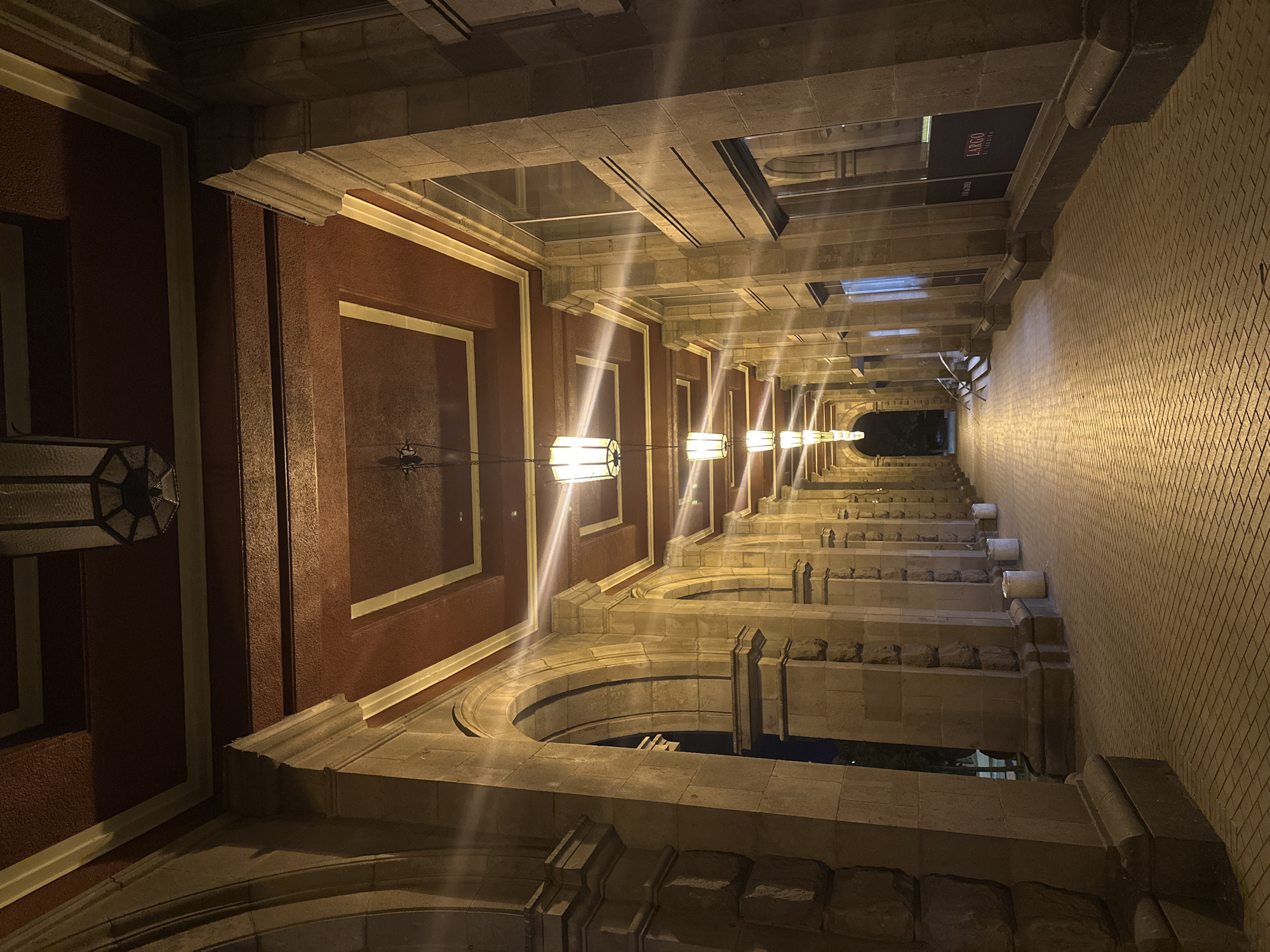 The image shows a long, arcaded walkway at night, illuminated by hanging lights and streetlights. The walkway is paved with cobblestones. On the right side of the walkway, there are large windows and a sign that reads "LARGO DE SERDICA". The architecture suggests a historic or classical style with stone columns and arches. The scene is likely in a public space, possibly a commercial area or a historical site in Sofia, Bulgaria, given the presence of the "LARGO DE SERDICA" sign, which refers to the archaeological complex in the city center. There are no people visible in the image. The lighting creates strong highlights and shadows, giving the scene a dramatic atmosphere.