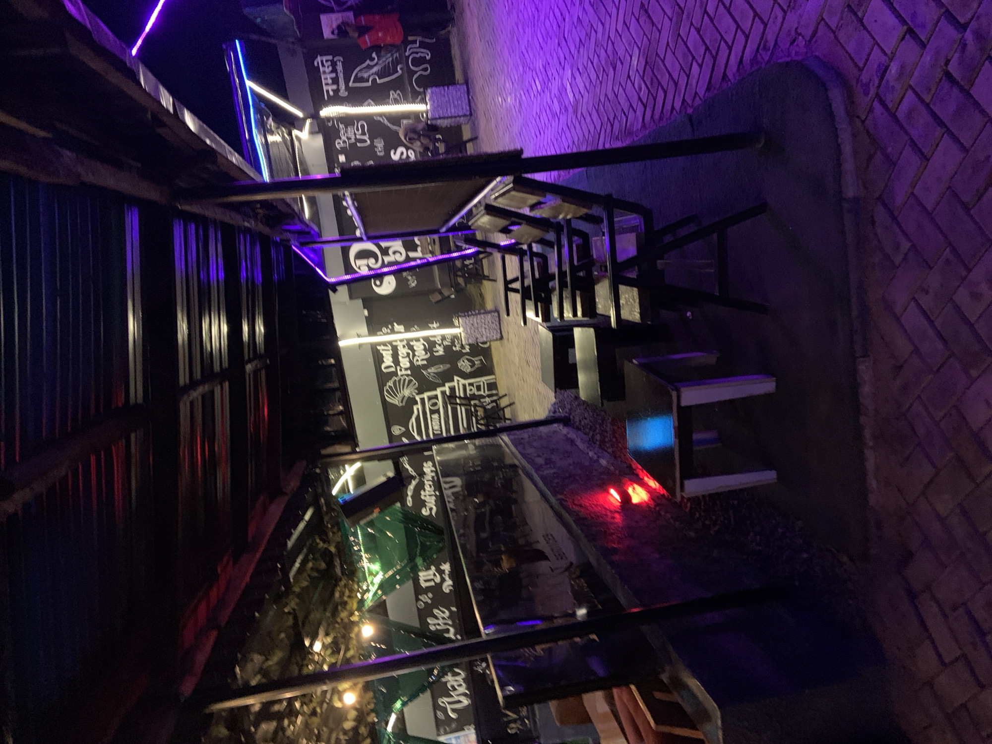This media file depicts an outdoor seating area at night, illuminated by vibrant purple and blue neon lights. The setting appears to be a restaurant or cafe, with tables and chairs arranged under a covered structure. The floor is paved with bricks, and the walls are adorned with black and white graffiti-style art. One section of the wall features text that includes "Namaste" and "Beer Us". Plants and string lights add to the ambiance. There are no people prominently featured, but the scene suggests a place where people would gather to eat and drink. The overall mood is lively and atmospheric, likely a popular spot for evening entertainment.