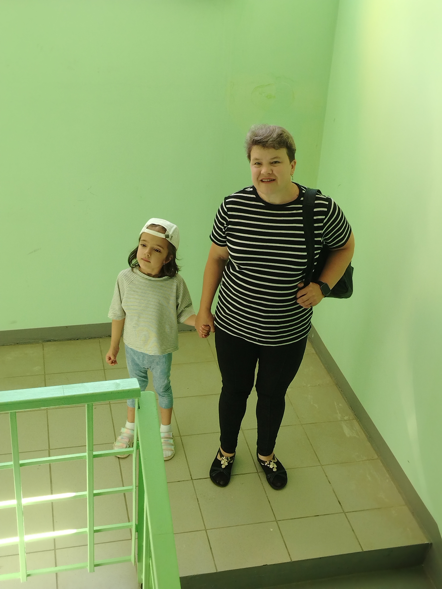 Two people, a woman and a young girl, are standing on a tiled staircase landing. The woman, on the right, is wearing a black and white striped t-shirt, black pants, and black flats adorned with a decorative bow and embellishments. She has a dark shoulder bag slung over her left shoulder and wears a smartwatch on her left wrist. Her short, grey hair is styled. The young girl, on the left, is wearing a horizontally striped t-shirt, light blue leggings, a white baseball cap, and light-colored sandals with multiple straps. She is holding the woman's right hand. They are both looking in different directions, with the girl looking slightly upwards and to her left. The environment is an indoor staircase with pale green walls and light-colored square tiles on the floor and steps. A green metal railing is visible to the left of the girl. The lighting appears to be artificial, casting long shadows from the railing. The location is identified as Noviy, Russia.