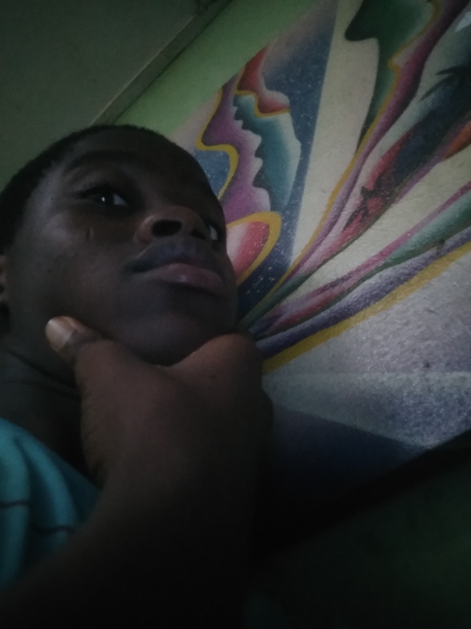 A person is resting their chin on their hand in a brightly-lit room. A colorful bedsheet with blue, pink, and yellow colors can be seen behind the person. The location in the city is Unknown, Unknown.