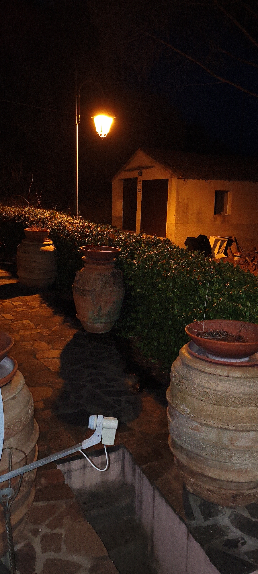 The image depicts an outdoor scene at night in what appears to be a rural or garden setting. A single lamppost with a warm, orange glow illuminates the area, casting long shadows. In the foreground, there are several large, weathered terracotta pots, some of which are empty and others containing sparse, dry plant matter. A surveillance camera is mounted on a metal arm, pointed towards the pots and the surrounding area. Behind the pots, a dense hedge forms a backdrop. Further back, a small, rustic building with a thatched roof and two large doors is visible, illuminated by the lamppost. The ground is paved with flagstones, some of which are wet and reflect the light. The overall impression is one of quiet, nighttime ambiance, possibly in an Italian countryside setting.