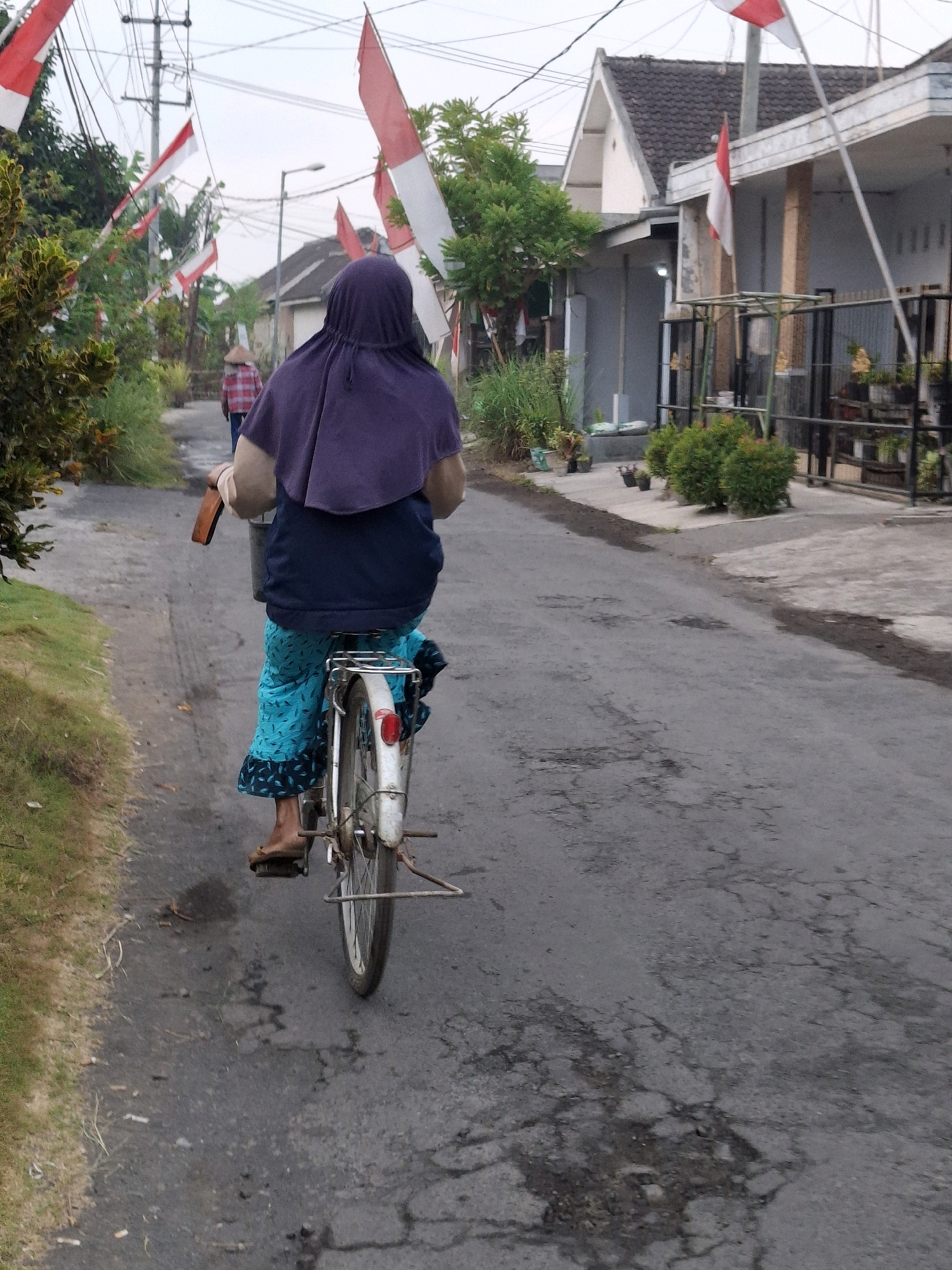 An elderly woman wearing a purple headscarf and a blue shirt rides a white bicycle down a cracked asphalt road. She is wearing a light blue patterned skirt and brown sandals. The bicycle has a basket on the back. The road is lined with trees and houses, some of which are decorated with Indonesian flags. In the background, a person wearing a conical hat walks away from the camera. The sky is overcast. This scene appears to be in a residential area in Lumajang, Indonesia, possibly during a celebration given the flags.