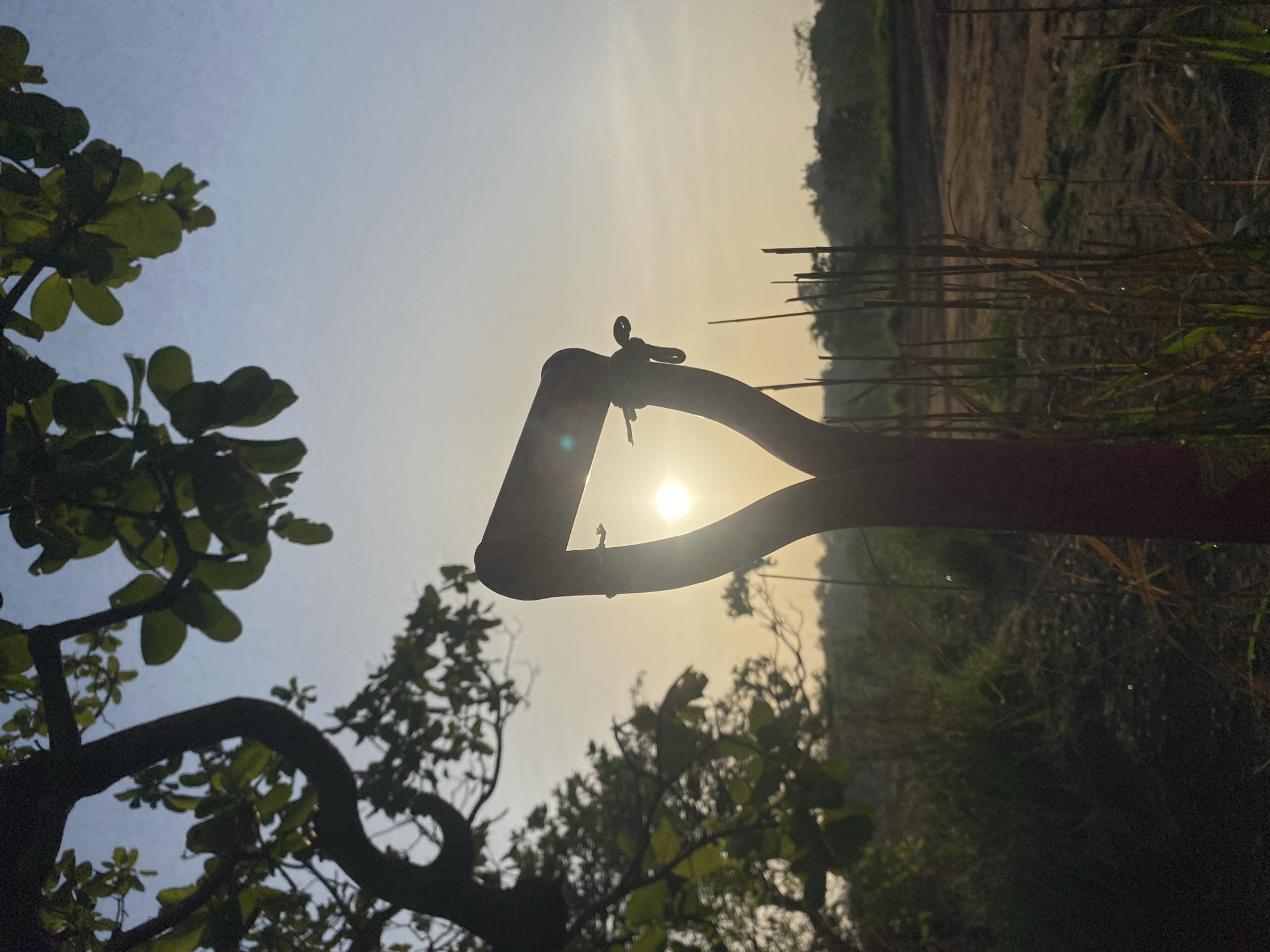 The image shows a shovel silhouetted against a bright, hazy sky with the sun visible through the handle. The setting appears to be outdoors, possibly a rural area. To the left, there are branches and leaves of a tree, while the background behind the shovel includes what appears to be vegetation and possibly a field. The lighting suggests either sunrise or sunset. Given the city of Amancho, Nigeria, is predominantly rural, the location likely lies within an agricultural region or outskirts of the city.