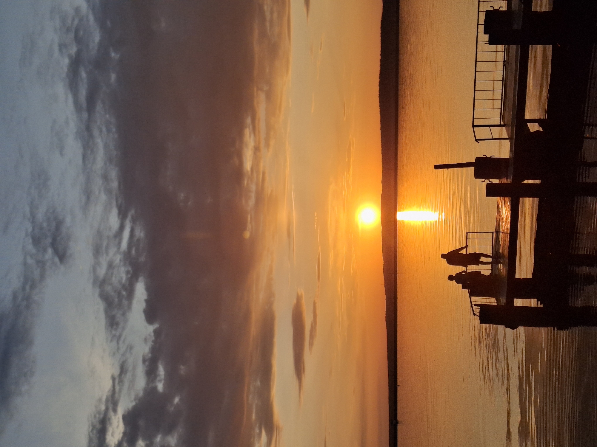 Here is a detailed description of the image:
The image captures a sunset over a body of water, likely a river or lake, near the city of Chaikovsky, Russia. The sky is filled with clouds, illuminated in shades of orange, yellow, and gray by the setting sun. The sun itself is a bright orb reflecting on the water, creating a shimmering path of light.
In the foreground, there is a silhouette of a pier or dock structure. On the dock, there are two figures, also in silhouette, appearing to be observing the sunset. The structure has railing details. The water is calm, reflecting the sky and the pier. The overall scene evokes a sense of serenity and peacefulness.