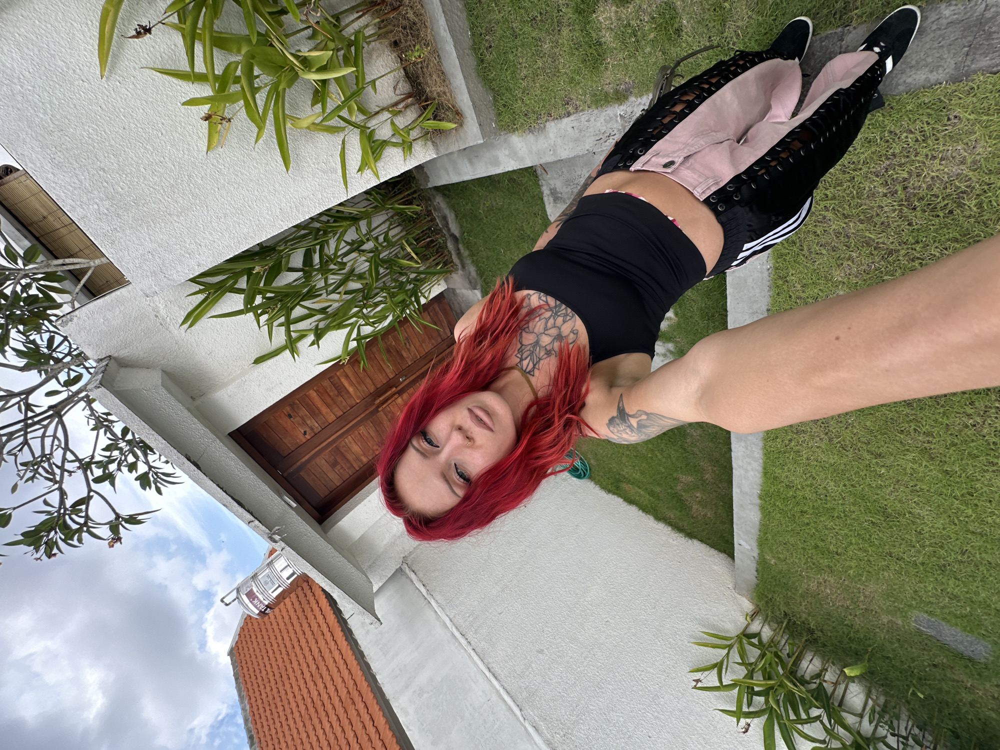 In North Kuta, Indonesia, a young woman with vibrant red hair and visible tattoos poses for a selfie outdoors. She wears a black tank top and distinctive pink and black lace-up pants, paired with black sneakers. The background reveals a white-walled building with a wooden door, a tiled roof, and lush green grass. A water tank is visible on the roof.