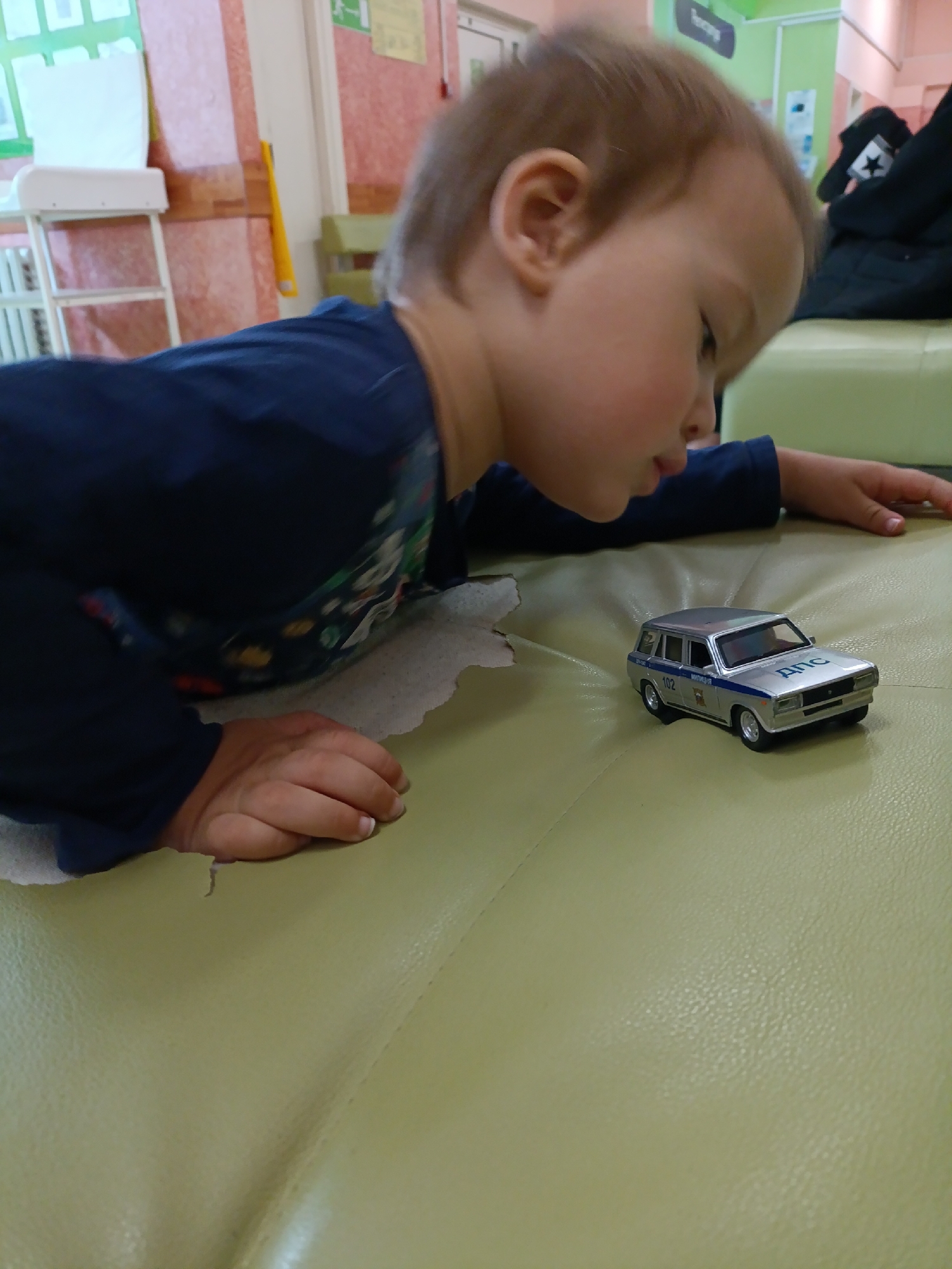 A young child in a blue long-sleeved shirt is lying prone on a green surface, possibly a sofa or a play area. The child's gaze is directed towards a silver toy car with "Police" or related markings on its side. The car is positioned on the green surface in front of the child. The location is difficult to determine from the image, and the assertion that it is in the city of Chaikovsky, Russia, cannot be confirmed from the visual information. There are other objects in the background, including what appears to be a changing table and a wall with signage.