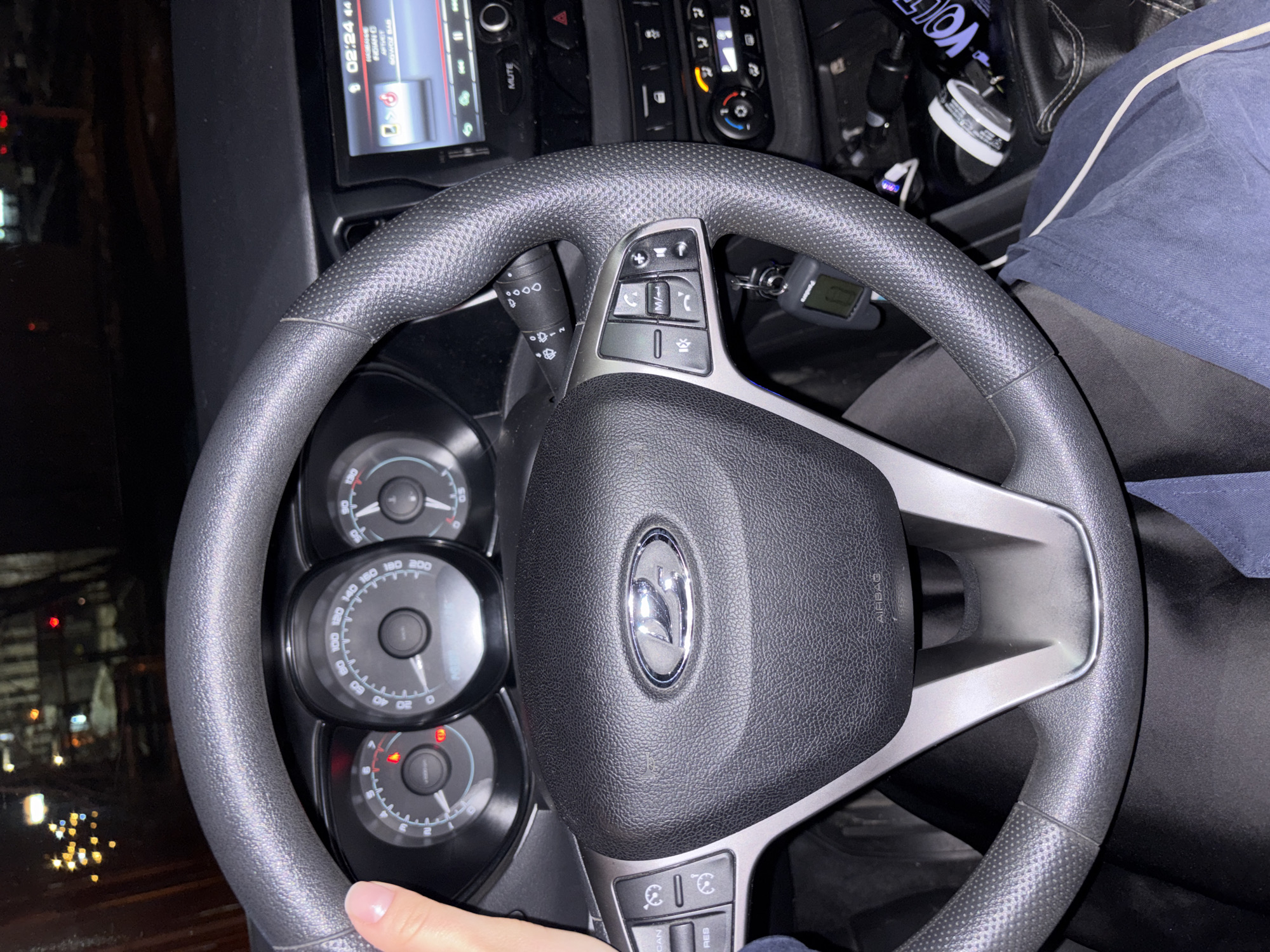 The image shows the steering wheel of a car, specifically a Lada Vesta, focusing on the steering wheel controls, dashboard, and part of the car's interior. The emblem on the steering wheel indicates it is a Lada. The dashboard displays gauges, including the speedometer and other indicators. The car appears to be in operation at night, as indicated by the interior lighting and visible streetlights through the windshield. A hand is visible on the steering wheel. A phone is visible in the upper part of the shot along with a can and a vape. Based on the 02:24 time on the dashboard, the time of day is early morning. Based on the can and the vape, the person is likely using those products. The OCR text "INDIAN O", "AFTVET" and "SOWOZ BAB" likely reference music playing.