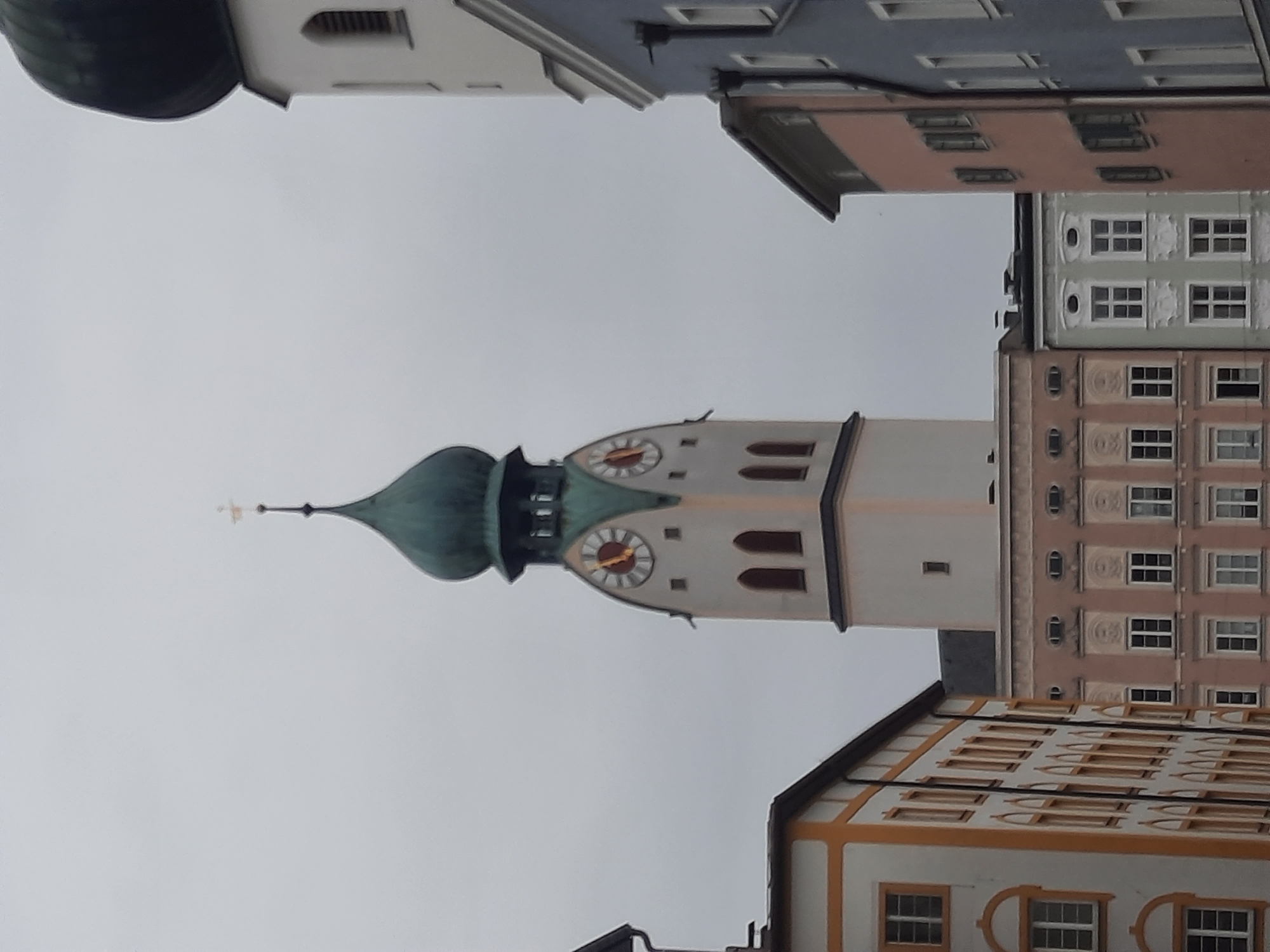 The image shows a view of a city street in Rosenheim, Germany, featuring several buildings and a prominent clock tower. The clock tower, which appears to be part of a church or historical building, has a green onion dome and two clock faces with roman numerals. The surrounding buildings are of various architectural styles and colors, including pastel pink, light blue, and white with orange trim. The sky is overcast and grey. The image is taken from a low angle, looking up at the structures. There are no people visible in the image.