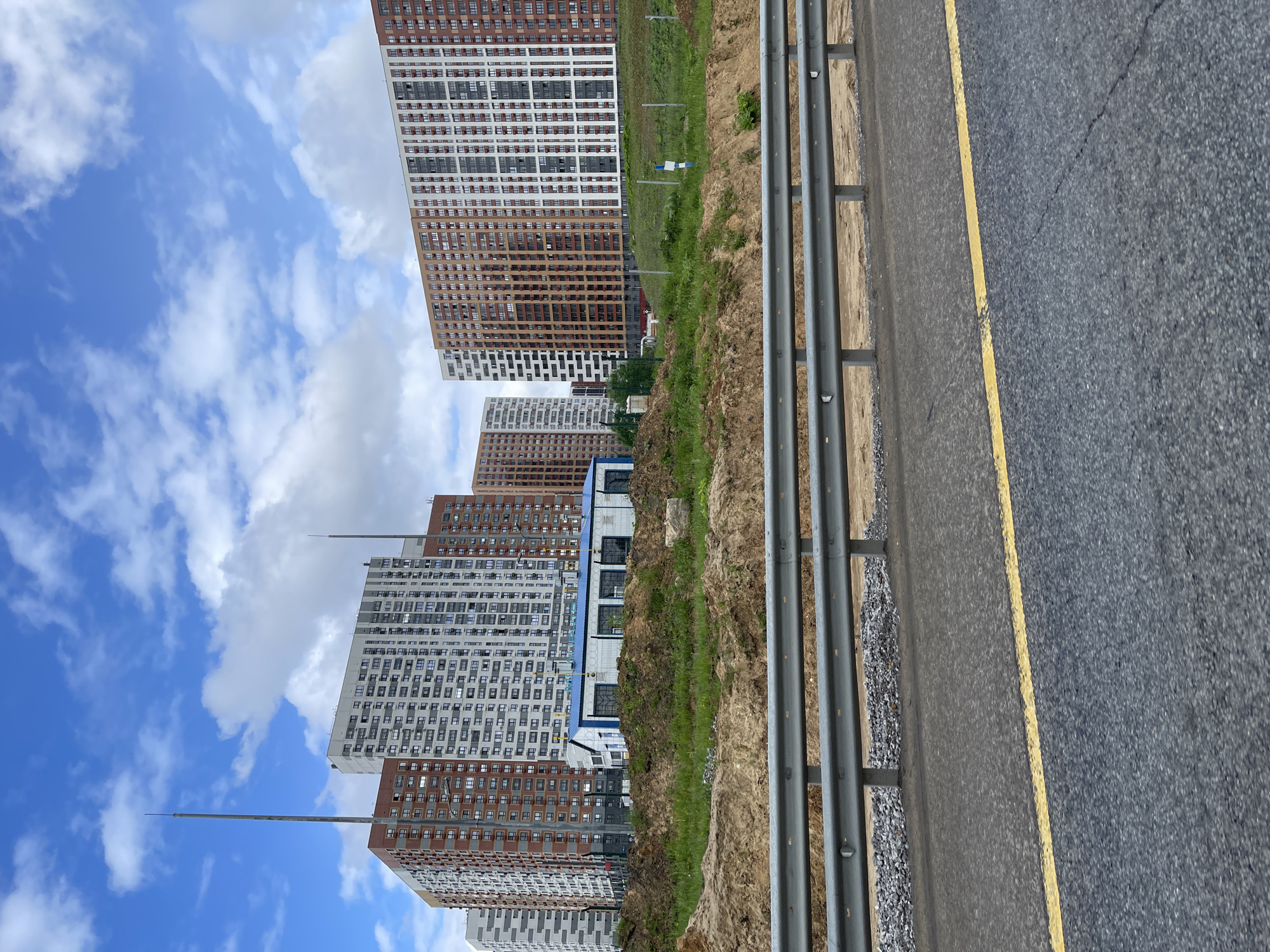 The image shows a view in Novodrozhzhino, Russia, featuring tall apartment buildings under a partly cloudy sky. A multi-lane highway with a yellow painted line on the road shoulder is visible on the right, separated from a grassy slope by a metal guardrail. The apartment buildings are primarily white and brown. One building has an antenna on top.