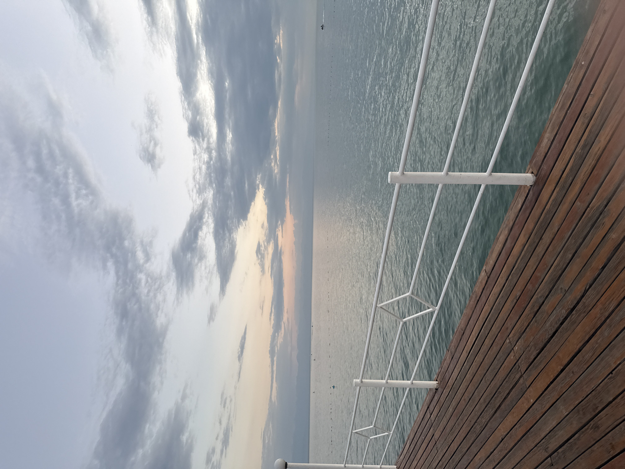 The image depicts a serene view of a body of water from a wooden pier in Sary-Oy, Kyrgyzstan. The sky is overcast with scattered clouds, tinged with the soft colors of dawn or dusk. The water is calm, reflecting the muted sky. In the distance, a thin line of mountains can be seen on the horizon. A white railing runs along the edge of the pier, with a repeating diamond pattern in its design. A few small buoys are visible on the water, and a single person is seen on a small boat in the far distance. The wooden planks of the pier are aged and weathered, adding a rustic charm to the scene.
