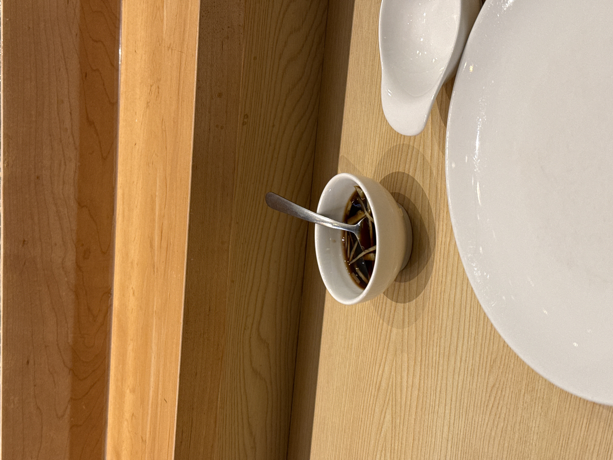 The image shows a wooden table with a white plate and a small bowl containing a dark liquid with thin noodles, a spoon is placed inside. The table is located next to a wooden structure, possibly a wall or furniture panel. The scene is indoors and likely in a restaurant. There is no indication of a specific location within New York City.