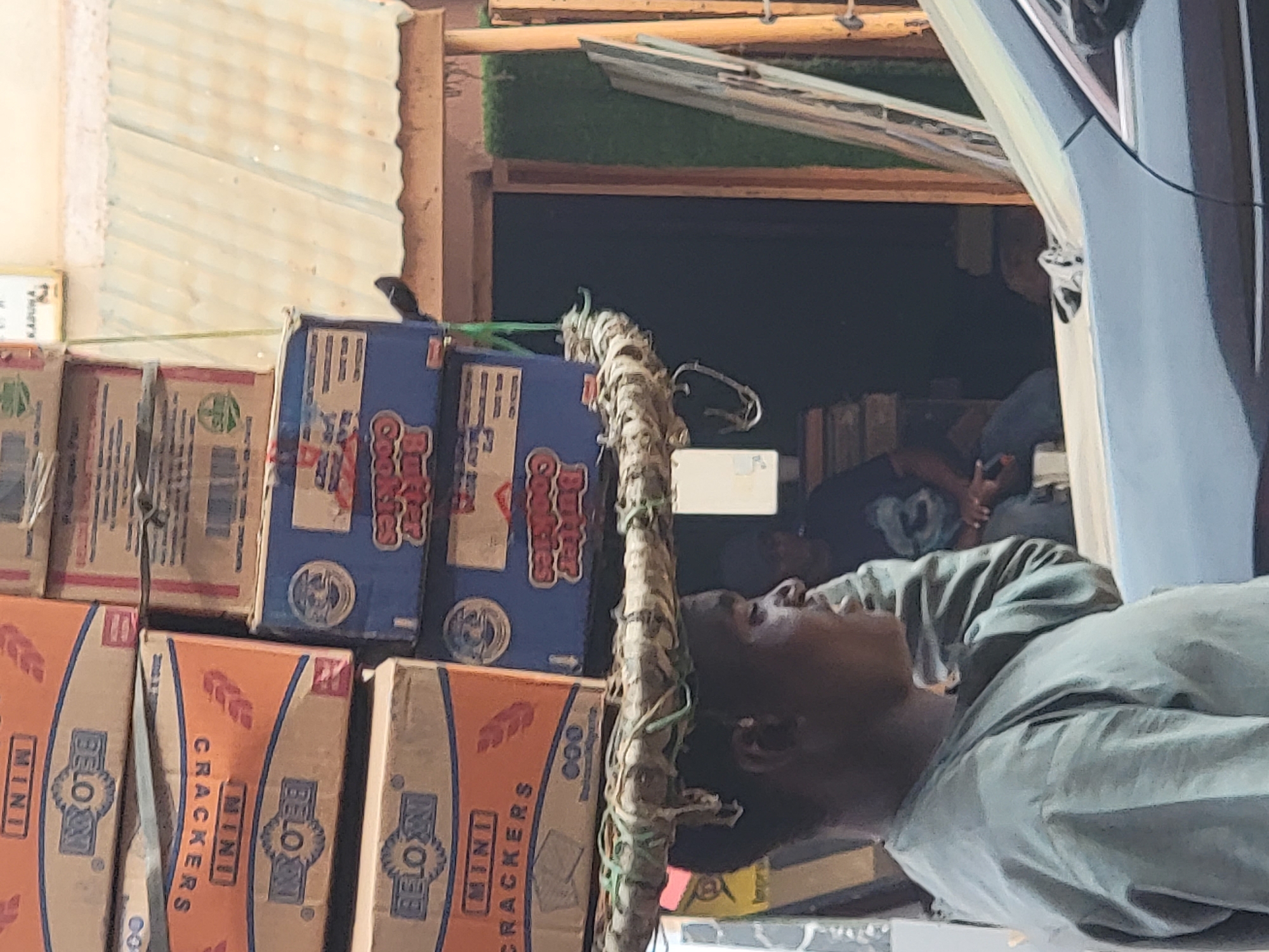A stack of cardboard boxes, printed with "BELOX MINI CRACKERS" and "Butter Cookies", is piled outside a shop in Maiburiji, Nigeria. A black bird is perched atop one of the boxes. A man wearing a light green shirt is seen near a vehicle. Inside the shop, there are at least two people seated.