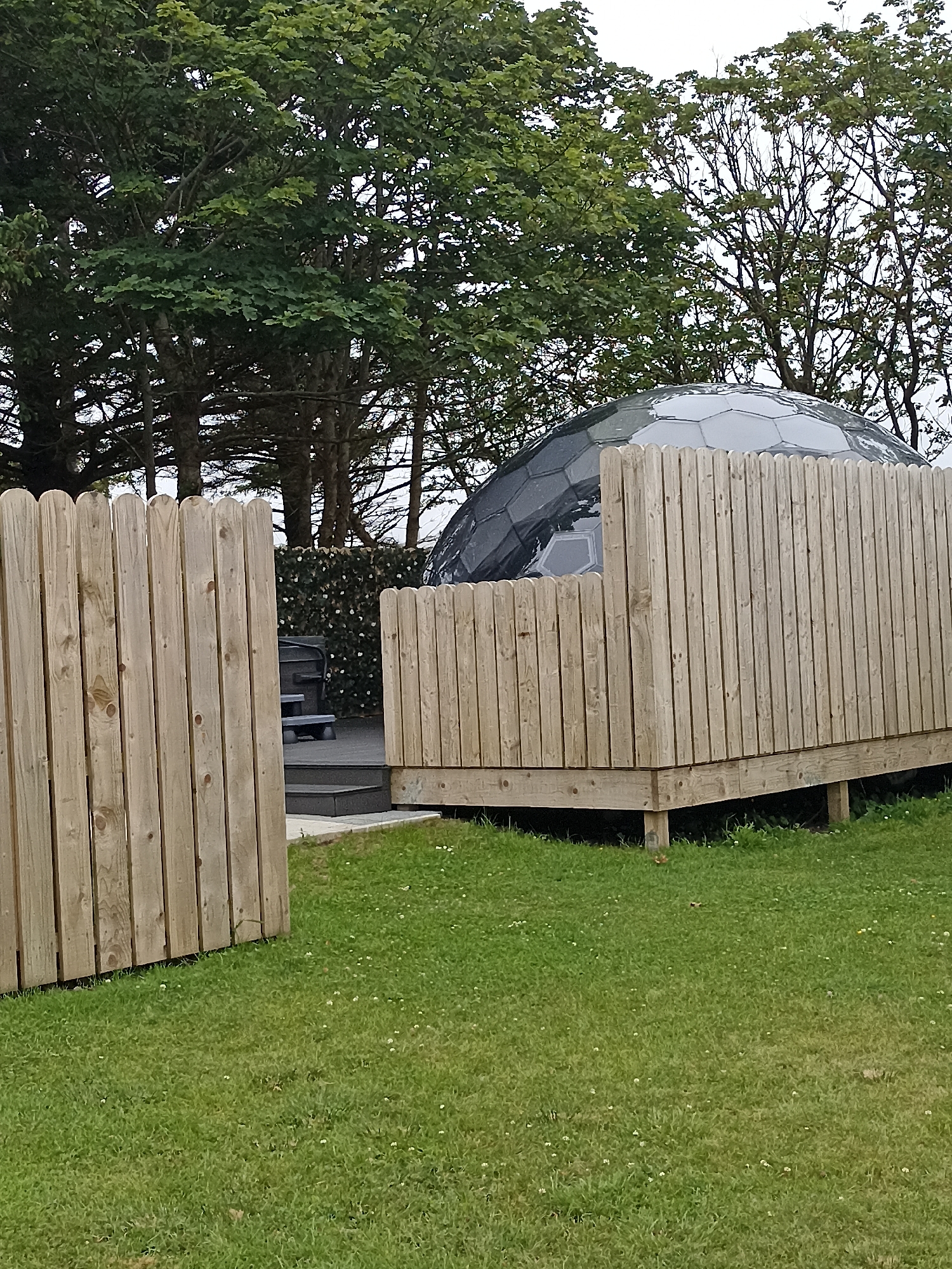 A geodesic dome, partially obscured by a wooden fence, is situated on a grassy area. The dome has a dark, faceted surface. A wooden deck surrounds the base of the dome, with a section of the deck extending forward and enclosed by the fence. The fence is constructed from vertical wooden planks with rounded tops. In the background, lush green trees form a dense canopy against a cloudy sky. To the left of the dome, a portion of a dark-colored hot tub or seating area is visible on a raised platform behind a fence. The overall scene appears to be an outdoor recreational or accommodation area.