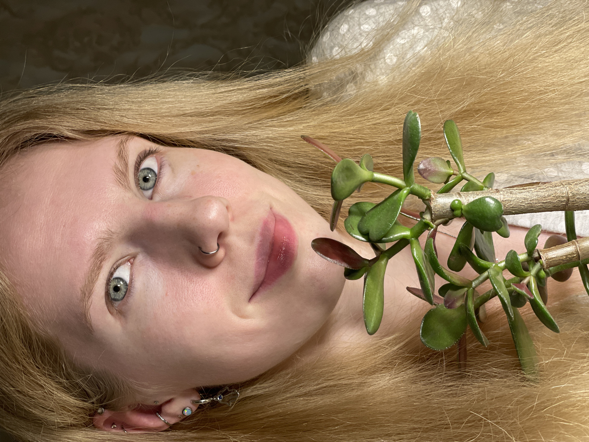 A fair-skinned person with blonde hair is lying down, facing the camera. The person has light blue eyes, a nose ring, and multiple earrings in their left ear. A jade plant lies on top of the person's hair. The background is a neutral color, possibly a wall. The location of Izhevsk, Russia is not evident in the image.