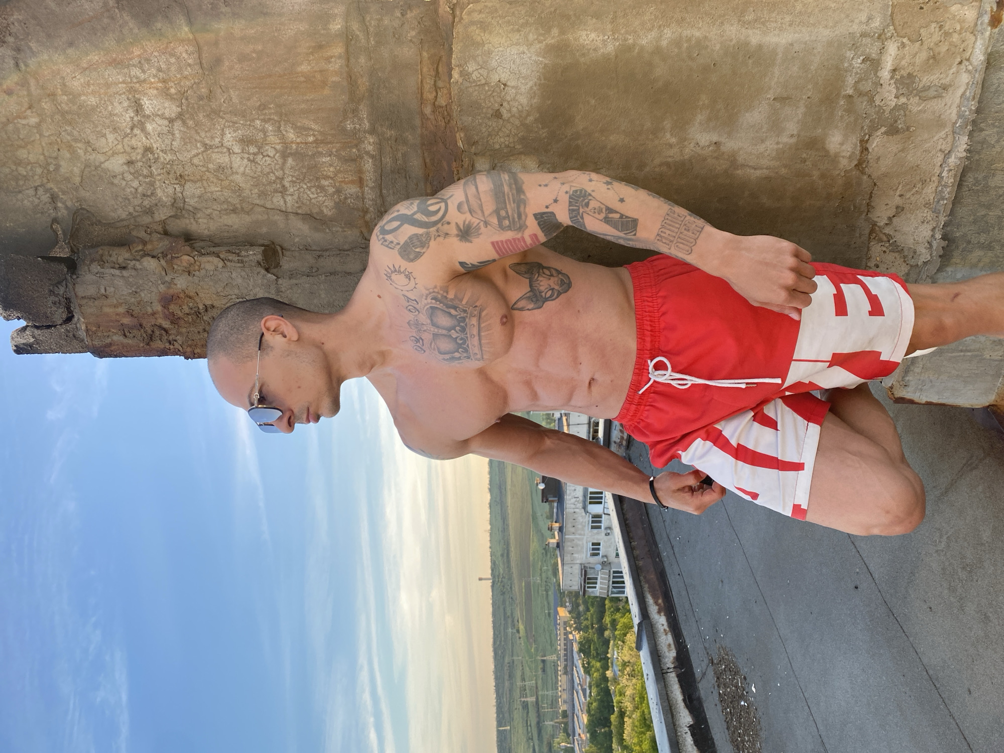 A shirtless man with tattoos stands on a rooftop edge in what appears to be Pleven, Bulgaria. He wears red and white shorts and sunglasses. The background shows a cityscape and a partially cloudy sky. The rooftop itself looks aged and cracked.