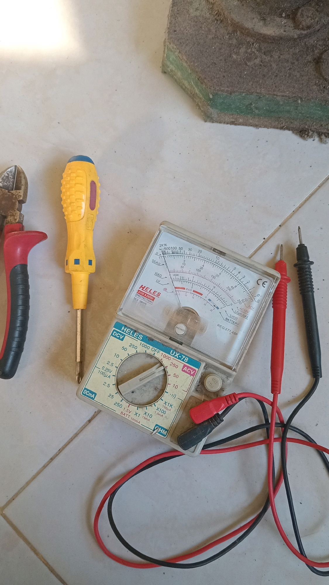 A close-up overhead shot shows a collection of electrical tools and a dirty, hexagonal-shaped object on a tiled floor. The tools include a pair of red and black wire cutters, a yellow and blue screwdriver, and a white analog multimeter with red and black probes attached. The multimeter has a dial that is slightly off-center, indicating it is not currently set to zero. The dirty object appears to be a piece of insulation or a sponge with layers of green and brown material. The lighting suggests an indoor setting, possibly a workshop or garage. The location is in the city of Randuagung, Indonesia.