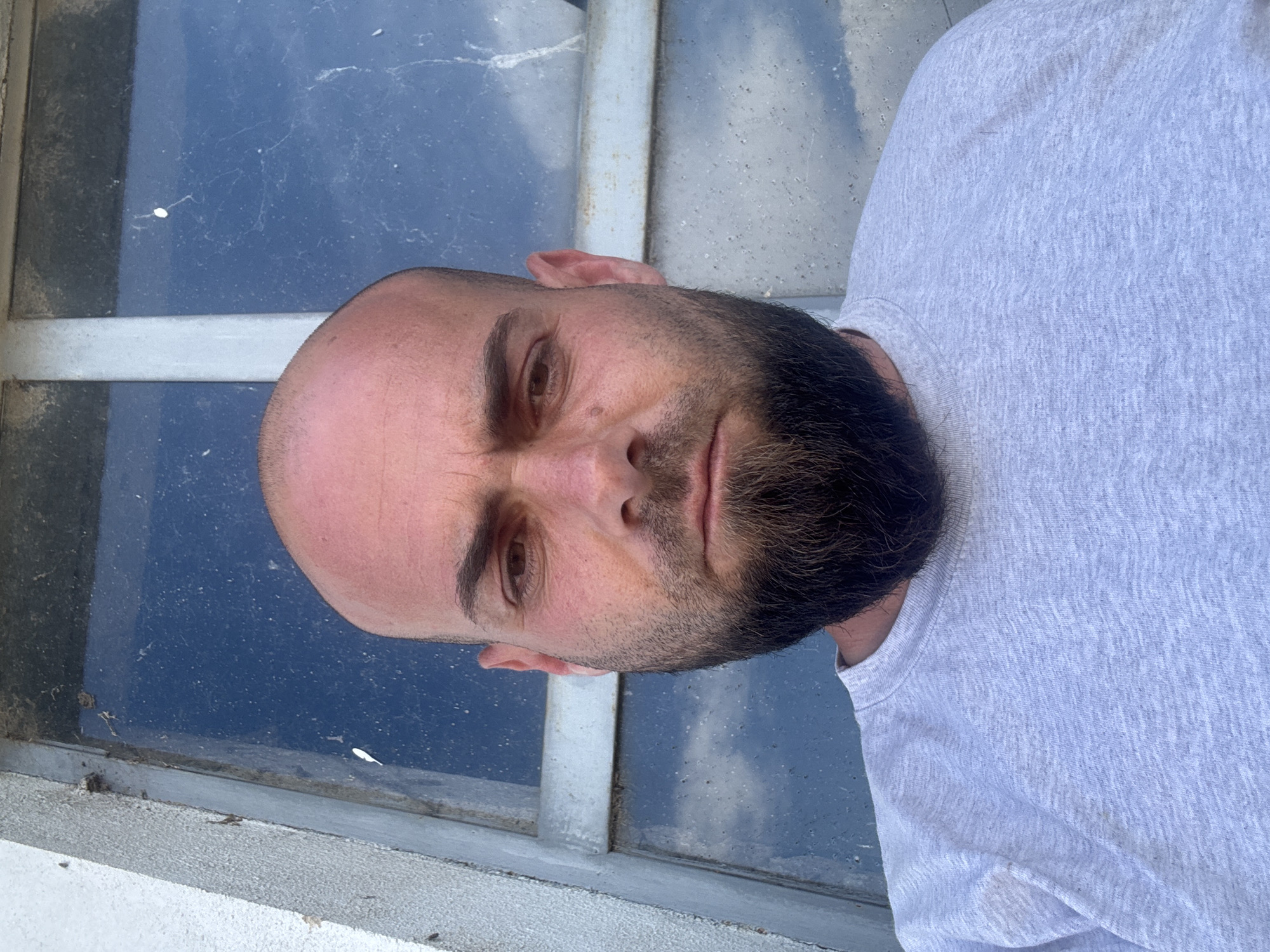 A medium shot depicts a bald, bearded man wearing a gray shirt, turned slightly to the side. He is positioned in front of a dirty, multi-paned window. The location appears to be Sarnevo, Bulgaria. The window is dusty and has cobwebs. The man’s expression is neutral. The exterior wall is weathered.