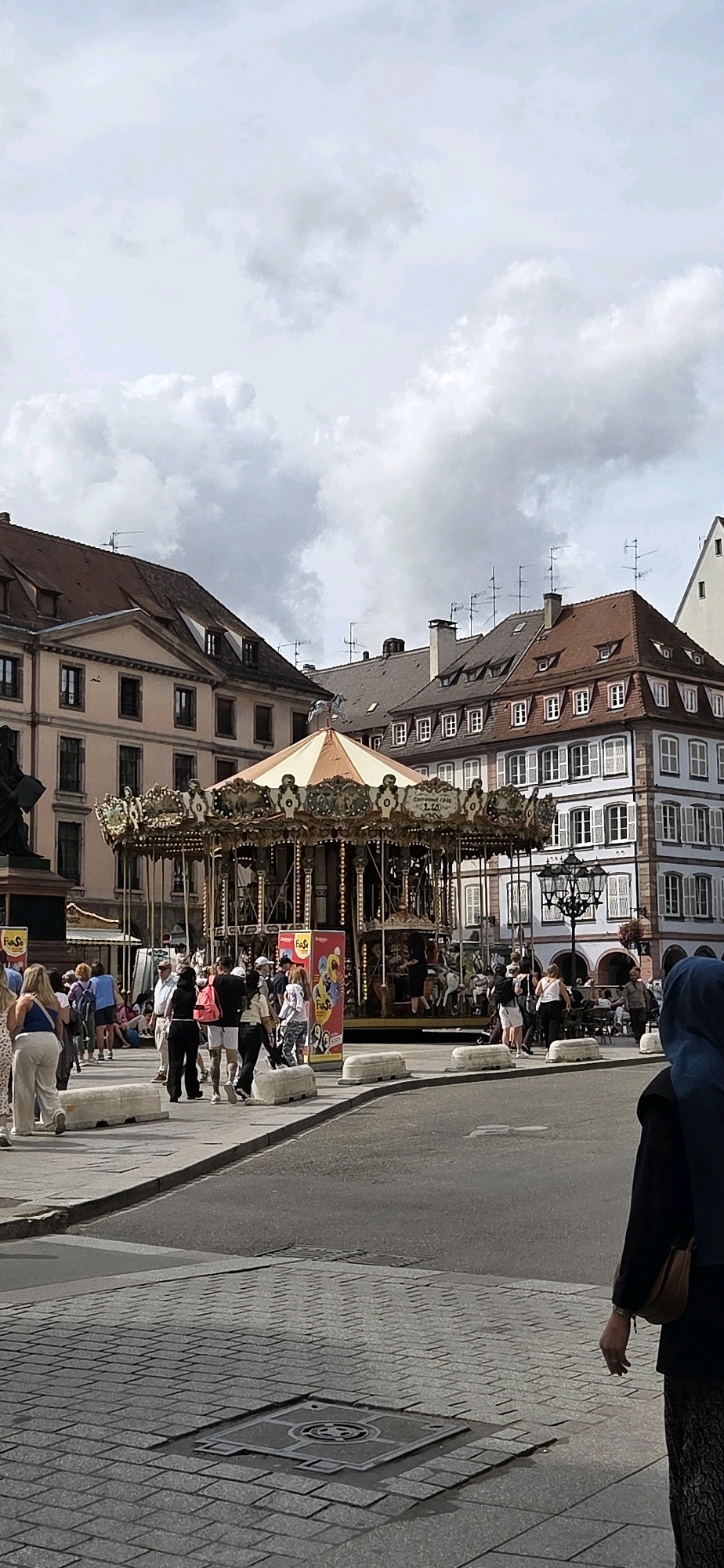 The image depicts a vibrant city square in Strasbourg, France, dominated by a classic carousel. The carousel, adorned with colorful horses and ornate decorations, is the central focus, with a crowd of people gathered around it. Children and adults can be seen interacting with the ride and enjoying the festive atmosphere.
The background features charming European architecture, with buildings showcasing distinctive Alsace-style facades. The buildings are a mix of pastel colors and traditional stone, with red-tiled roofs and numerous windows. Some buildings display architectural details like shutters and balconies. A few trees and streetlights add to the urban landscape.
The sky is partly cloudy, with dramatic, textured clouds that suggest either an approaching storm or the aftermath of one. The lighting suggests it is daytime, possibly late afternoon, with some shadows visible on the ground.
In the foreground, cobblestone paving and a street are visible, indicating a public area. Several people are walking through the square, some casually dressed and others appearing to be tourists. One individual in the lower right corner, wearing a dark top and a headscarf, is partially visible, with a brown bag slung over their shoulder. The overall scene conveys a sense of lively public life and leisure within a historic European city.