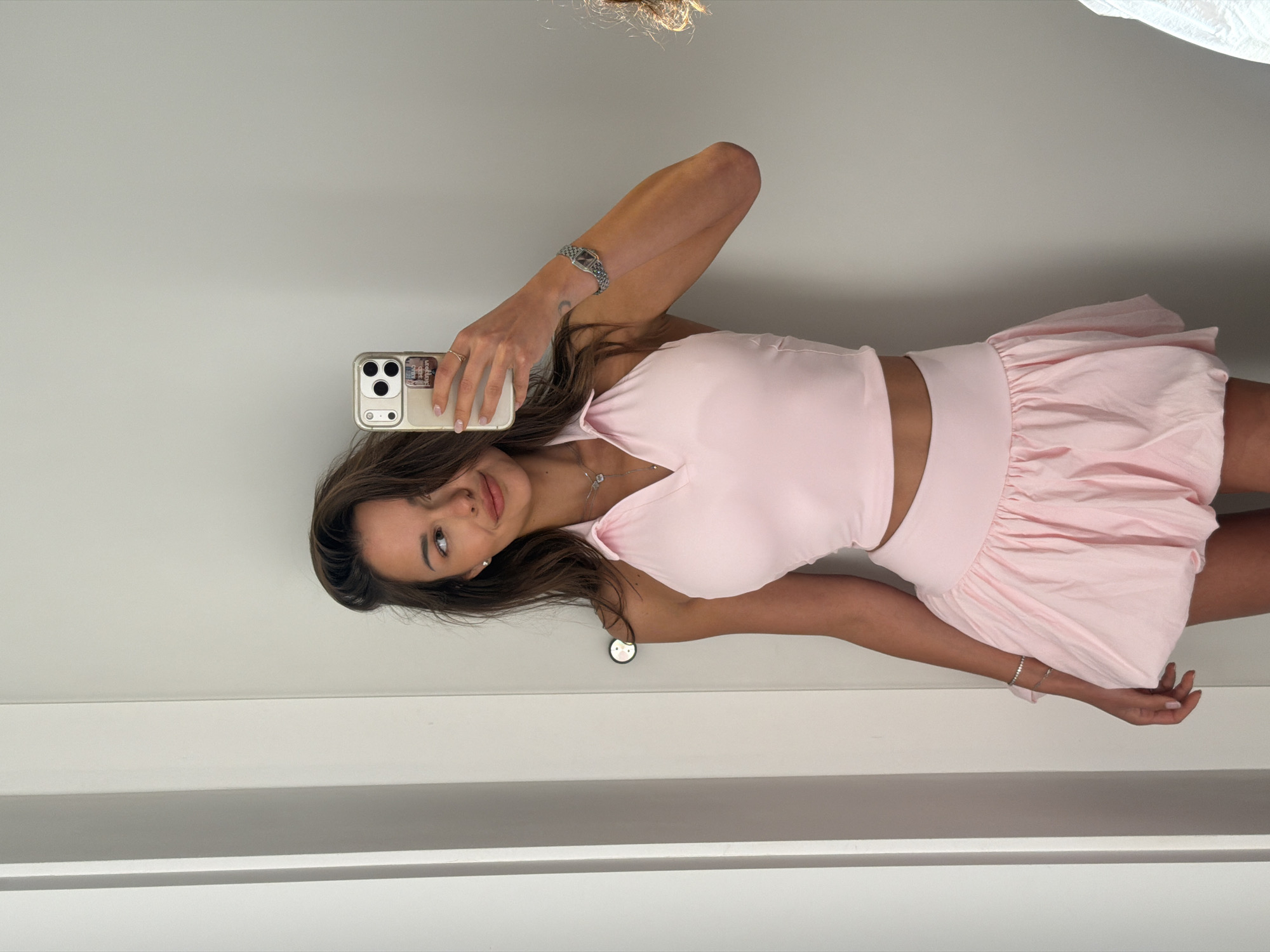 A young woman with long, dark brown hair and fair skin is taking a mirror selfie in a room. She is wearing a light pink halter-neck crop top with a collared neckline and a matching light pink bubble skirt. She is also wearing a silver necklace with a pendant, a silver bracelet, and a silver watch on her left wrist. Her right hand is holding a white smartphone with a gold case and a clear screen protector. The background appears to be a closet or dressing room, with a white door on the left and a grey wall behind her. There is no indication of the city of Saint Petersburg, Russia in the image.