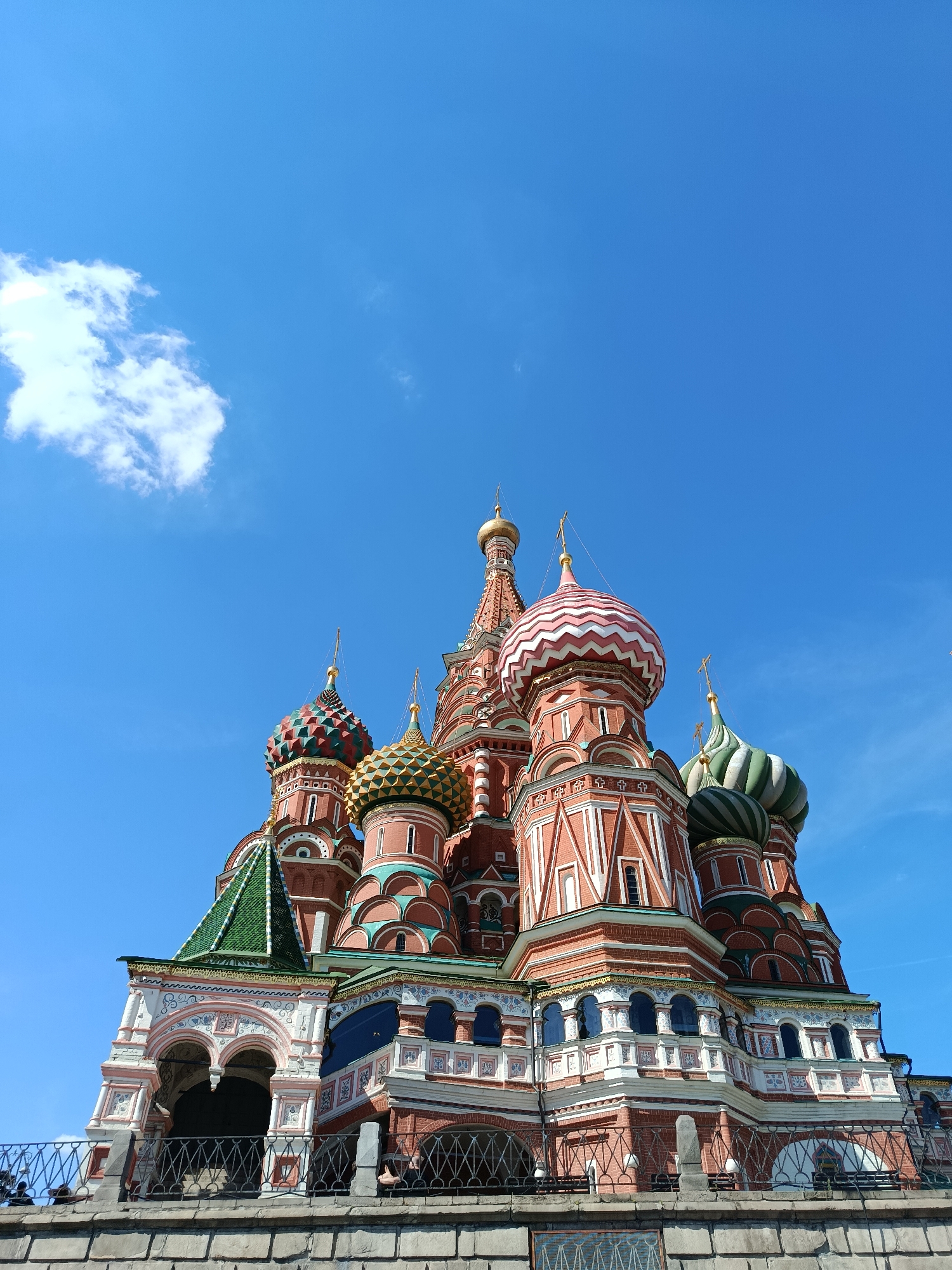 This is a low-angle, full-frame shot of Saint Basil's Cathedral in Moscow, Russia, on a clear day. The cathedral's iconic colorful onion domes and intricate architecture dominate the frame, set against a vibrant blue sky with a single, fluffy white cloud in the upper left corner. The lower portion of the image shows a stone wall and a metal fence, with glimpses of what appear to be people behind the fence. The sun is casting shadows on the building, indicating it's likely daytime.