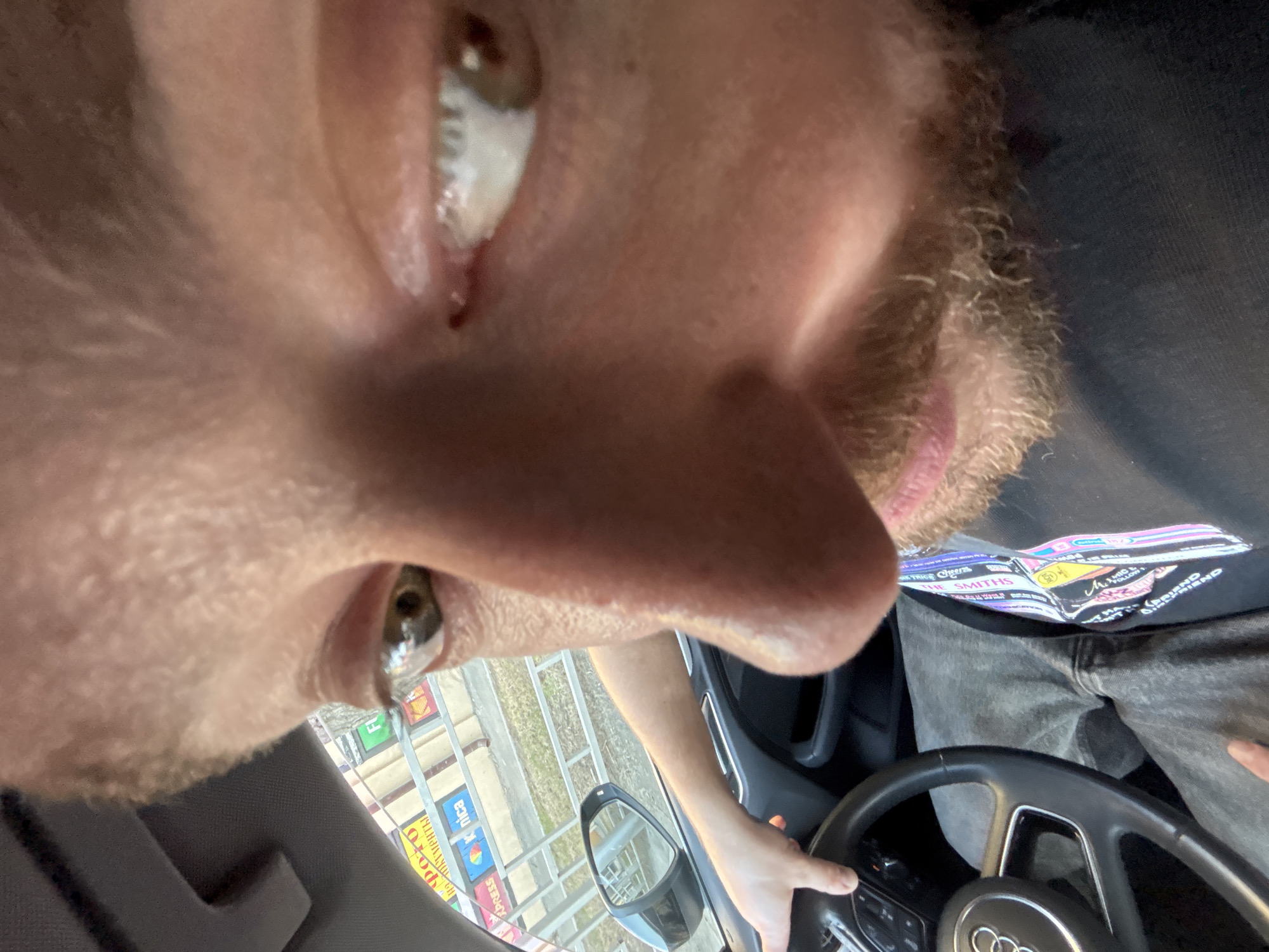 This image offers an unusual, extreme low-angle perspective of a person, likely male due to the visible beard and stubble, sitting in the driver's seat of a car. The camera is positioned directly below the face, capturing a close-up of their eyes, nose, and upper part of their mouth, with the eyes looking directly downwards towards the camera.
The setting is the interior of a car, indicated by the dashboard and a prominent Audi-branded steering wheel, which the person's right hand is resting on. The individual is wearing a black t-shirt adorned with a colorful graphic print that includes the text "THE SMITHS," paired with grey jeans.
Through the car's windshield and passenger-side mirror, an outdoor environment is visible under what appears to be clear daylight. The scene outside includes paved ground, some greenery, and various structures or fencing. Crucially, several colorful billboards and signs are visible, displaying Cyrillic text. Partially legible words include "ника" (nika), "фото" (photo), "экспресс" (express), and "реклама" (reklama - advertisement), which strongly align with the given location context of Krasnoyarsk, Russia.
The activity suggests the person is either driving, has just stopped, or is about to commence driving, while also engaging in taking a selfie from an unconventional angle. The expression is neutral, primarily focused on the act of looking at the camera.