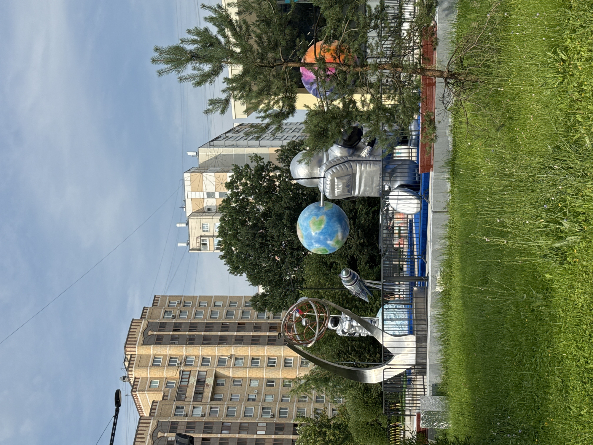 The image shows an outdoor sculpture park with a space theme in the city of Zelenograd, Russia. In the foreground, there is a patch of green grass with some wildflowers and a small pine tree. Behind the grass, a black metal fence encloses a paved area where the sculptures are displayed. The main sculpture features a silver-colored astronaut figure holding a globe of Earth. To the left of the astronaut, there is a metal structure resembling a stylized rocket or satellite dish, with a smaller astronaut figure inside and a model of the solar system. To the right of the main astronaut, another globe, this one brightly colored with purple and orange hues, is visible. In the background, there are tall, modern apartment buildings, typical of Soviet-era architecture, and lush green trees. The sky is partly cloudy. The overall scene depicts a public art installation celebrating space exploration, situated within a residential urban environment.