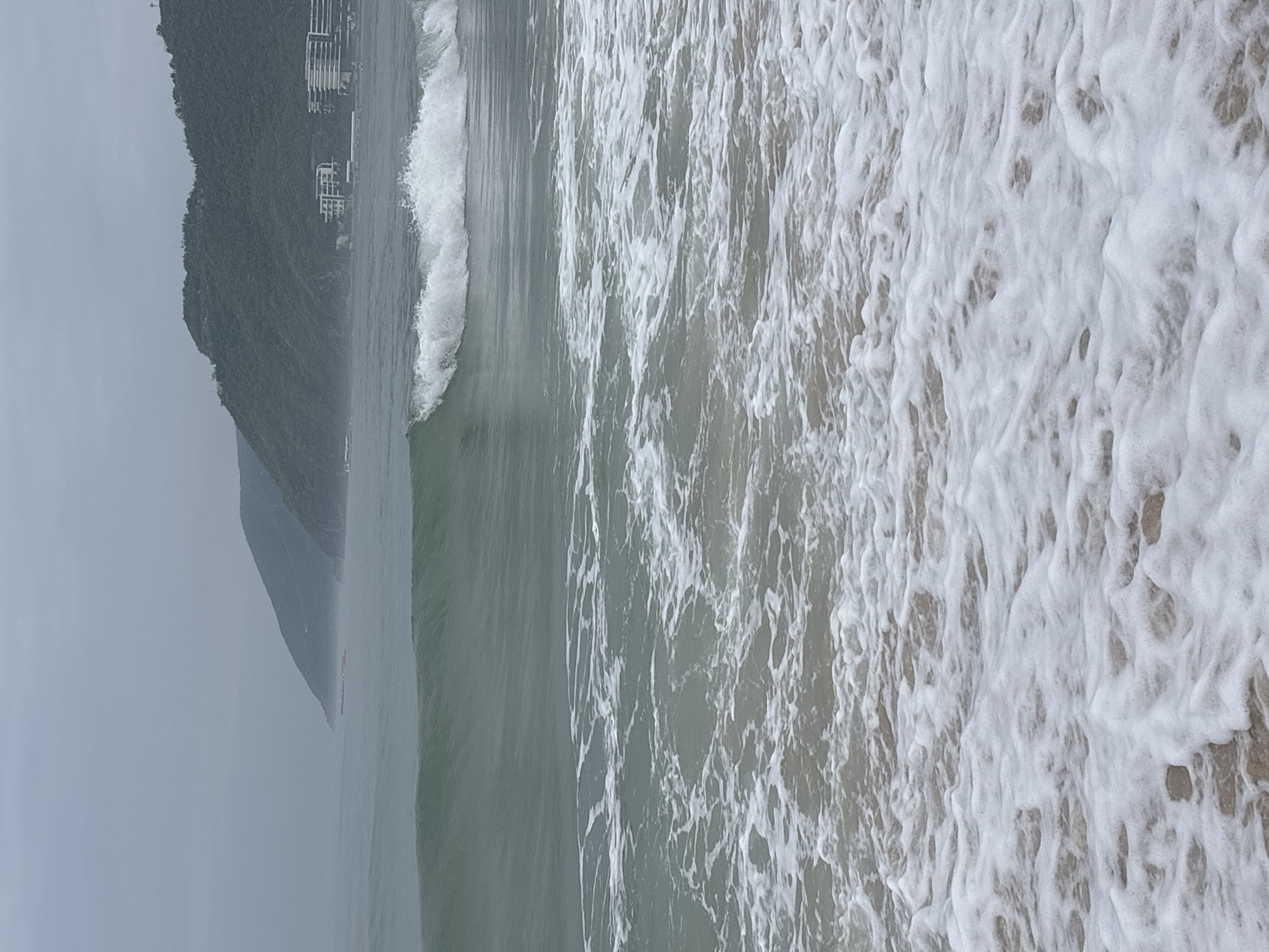 The image depicts a beach scene with waves rolling in. In the foreground, the water is foamy as it washes onto the sandy shore. A moderately sized wave is breaking in the mid-ground. In the background, lush green hills rise up, with a few buildings visible on the slopes and along the coastline. The sky is overcast and a hazy mist hangs over the distant landscape. The location is identified as a beach in Jiyang District, China. There are no people visible in the image.