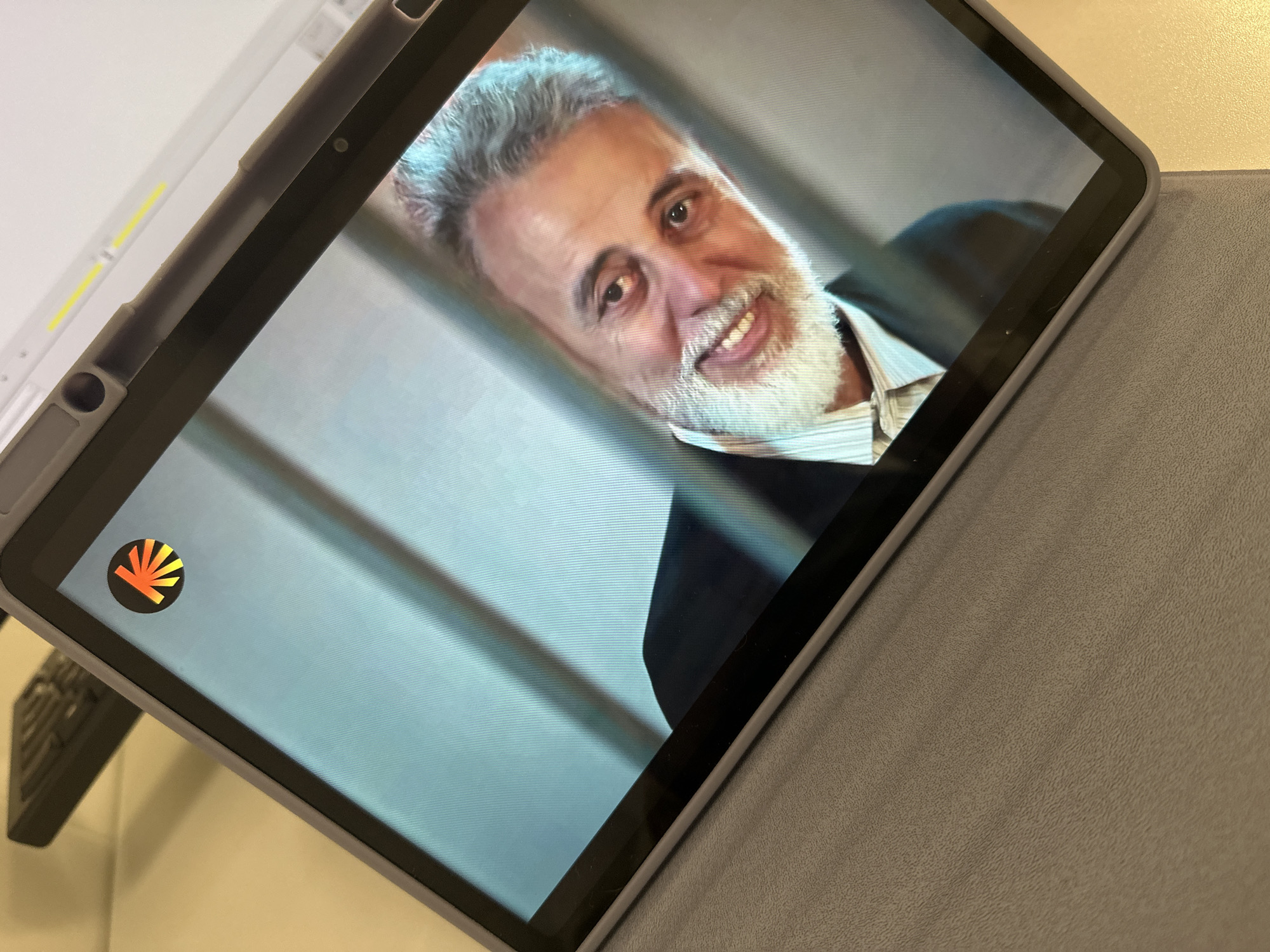A tablet displays a man with grey hair and a white beard, wearing a light-colored striped shirt and a dark suit jacket. The tablet is in a grey case and rests on a light-colored surface. A logo with a black circle and orange radiating lines is visible on the screen in the lower left corner. The location of the image is not discernible from the visible contents.