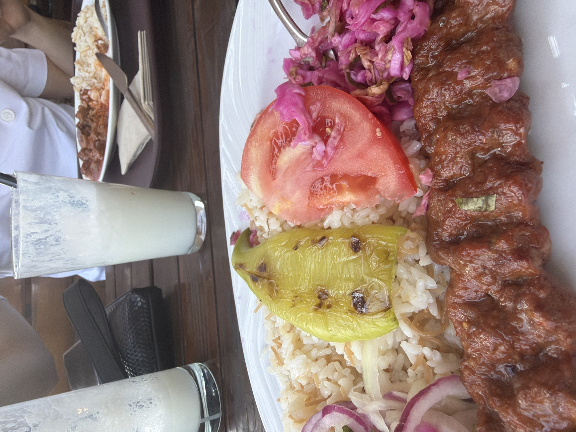 A close-up, eye-level shot shows a plate of food on a wooden table, possibly in Sofia, Bulgaria. The plate contains a kebab skewer, rice, a grilled green pepper, a slice of tomato, and a salad of shredded red cabbage and possibly onions. Two glasses of a white beverage, likely yogurt drinks, are visible in the background, along with a wallet and part of a person's arm. The setting appears to be outdoors, with a blurred background that suggests a casual dining environment.