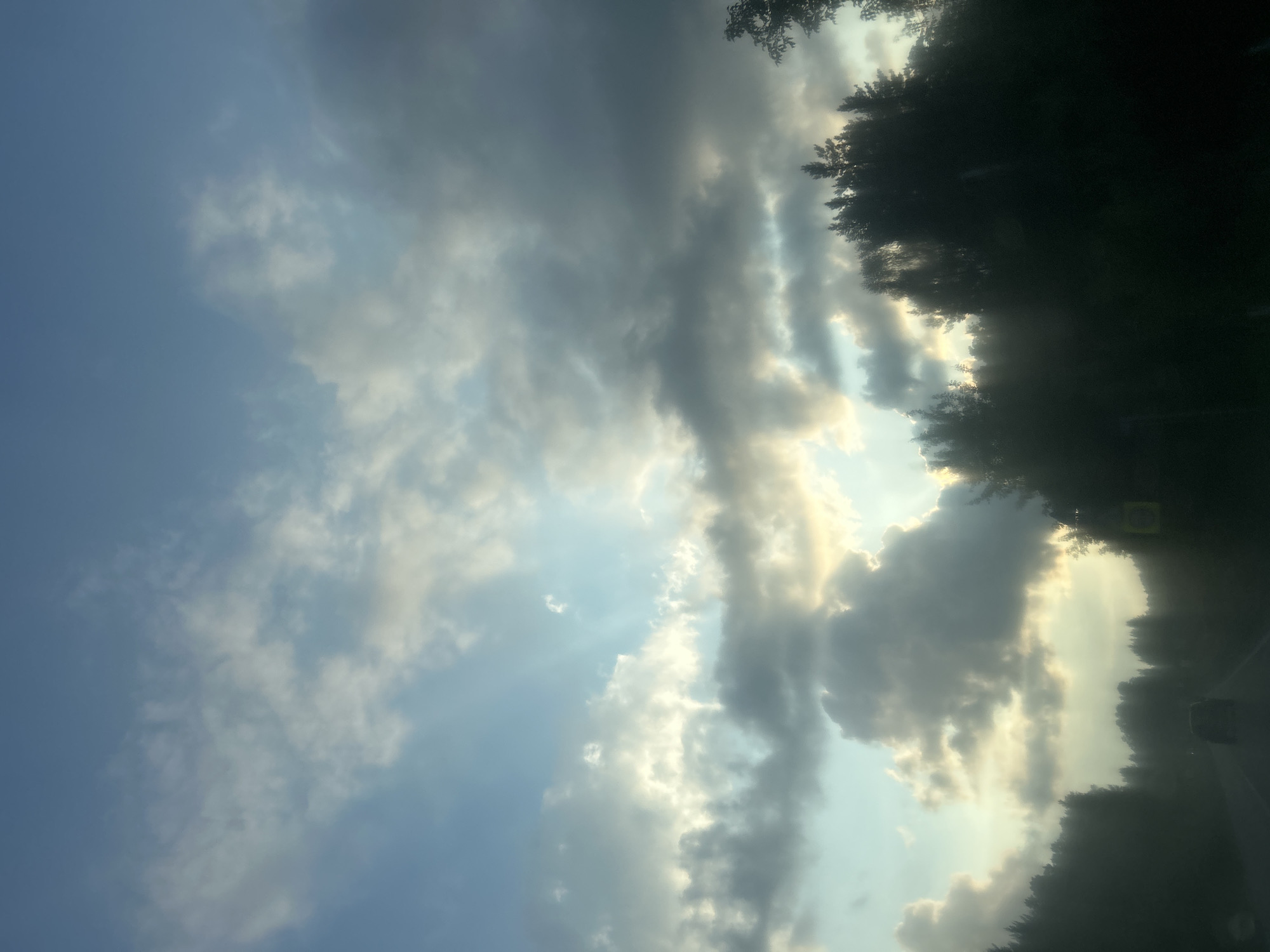 The image captures a view from a vehicle moving along a road lined with dense trees. The sky is filled with dramatic clouds, with rays of sunlight breaking through in several places, suggesting either sunrise or sunset. The overall lighting is dim, with the foreground and the trees appearing as dark silhouettes against the brighter sky. A single car is visible ahead on the road. The scene appears to be set in the city of Kurganovo, Russia, based on the context provided.