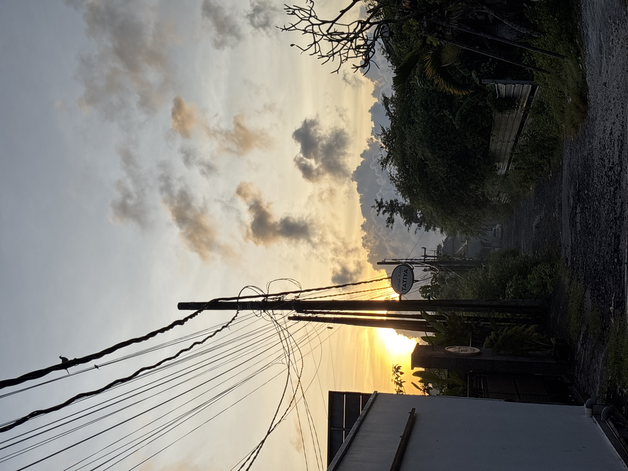 A street scene in Kuta Selatan, Indonesia at sunset. The sky is filled with clouds, ranging from light gray to dark gray. The sun is low on the horizon, casting a warm orange glow through the gaps between buildings and foliage. Power lines crisscross the sky in the foreground. Vegetation is abundant on the side of the road, with various trees and plants visible. A sign, "Rumah Kayu Tatiya", hangs from a pole. A section of a building is visible at the bottom of the frame. The road appears to be wet.