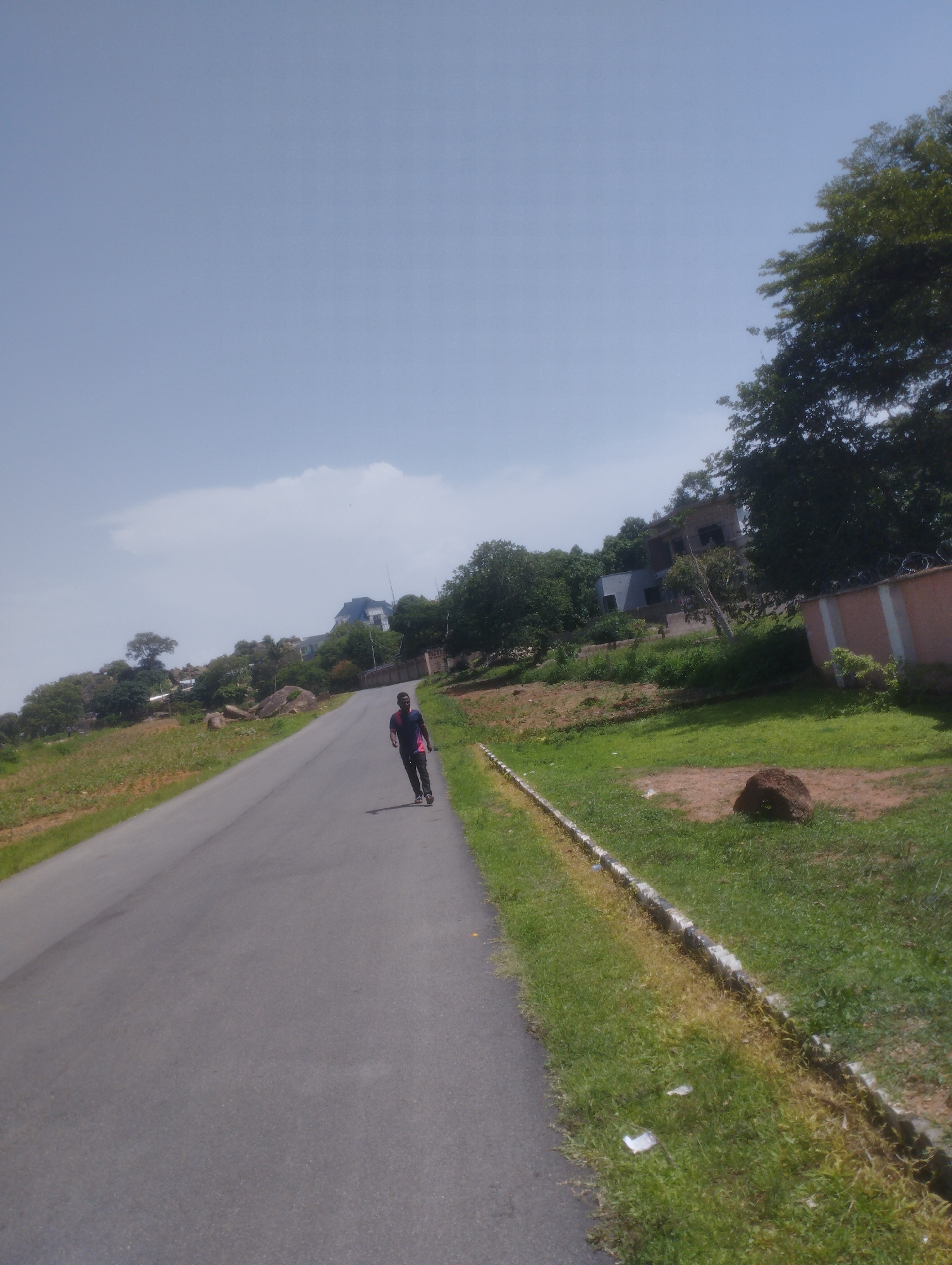 A person is skateboarding down a paved road in a somewhat hilly, semi-urban area. The road is asphalt and curves to the left as it goes uphill. On the left side of the road, there is a grassy slope with some rocks and scattered vegetation. On the right side of the road, there is a well-maintained lawn bordered by a concrete curb and then a long, light-colored wall topped with barbed wire. Behind the wall, there are trees and buildings, including a modern, multi-story house. The sky is mostly clear with a few wispy clouds. The scene appears to be in a suburban or semi-rural part of Jos, Nigeria, likely on a sunny day.