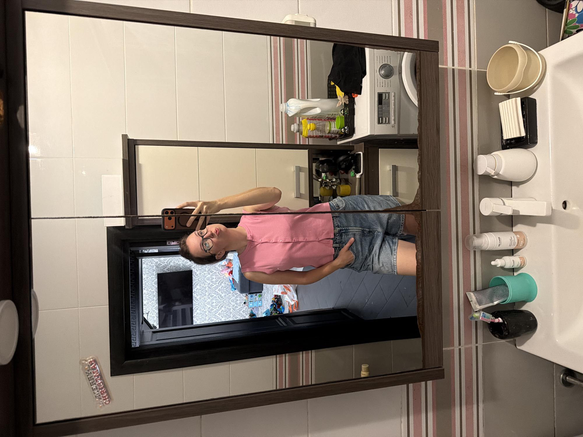 A woman is taking a mirror selfie in a bathroom. She is wearing a pink sleeveless top and denim shorts. In the reflection, a washing machine and several bottles of cleaning supplies are visible. The bathroom walls are tiled with a striped pattern. On the sink counter, there are toiletries such as toothbrushes, toothpaste, and soap. The overall environment appears to be a domestic bathroom.