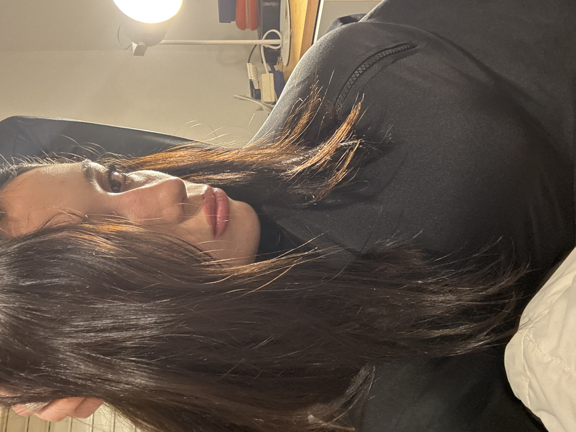 A woman with long, dark brown hair lies sideways against a light-colored pillow. She is wearing a black zip-up garment. Overhead, a lamp casts a bright light. Background details include power outlets and cables mounted on a wall. The location cannot be determined.