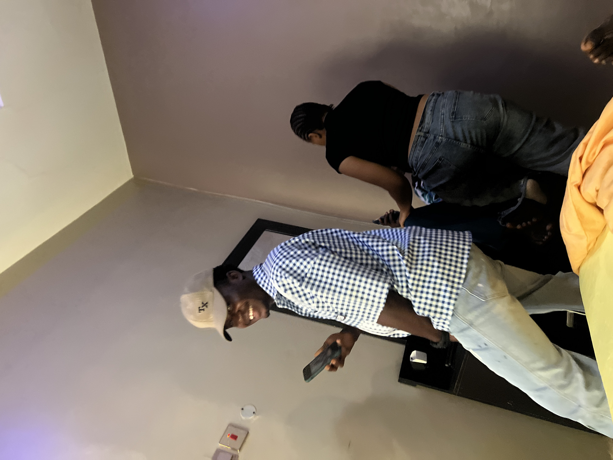 In an indoor setting, possibly a room in Nkpor, Nigeria, a man wearing a plaid shirt and a baseball cap stands holding a mobile phone. He is looking to his left and smiling. Behind him, a woman in a black top and jeans is seen from the back, leaning against a wall or furniture. The room has a plain wall and what appears to be a bed or couch covered with an orange blanket in the foreground. There are also some objects on the wall that appear to be light fixtures or switches. The lighting in the room is dim and appears to be artificial.