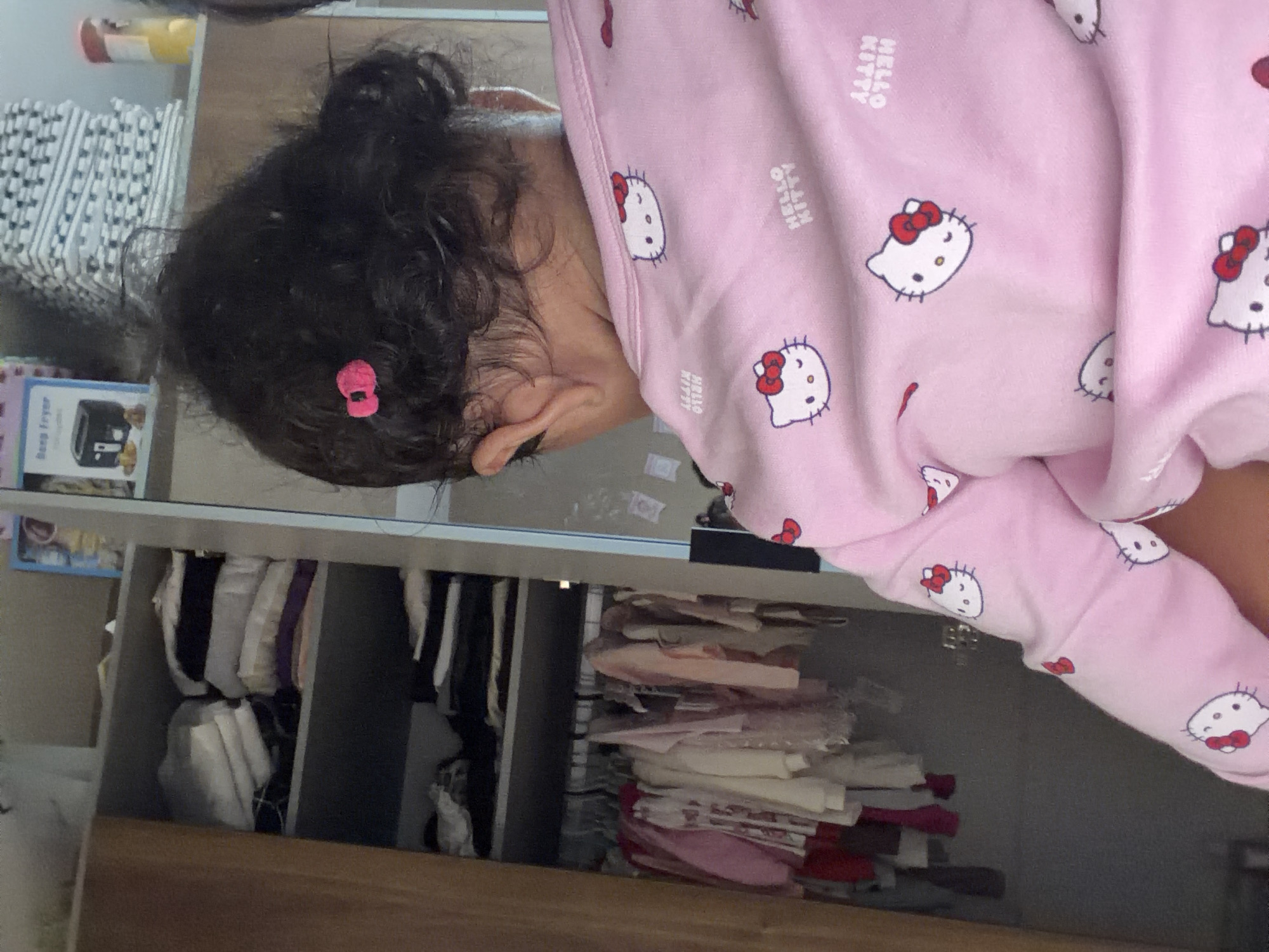 The image shows a young child from behind, wearing a pink Hello Kitty pajama set. The child has dark, curly hair pulled up with a pink hair clip. To the left, a wardrobe is visible with shelves containing folded clothes and hanging garments. On the right, a shelf holds a small appliance box and stacked items. The scene appears to be indoors, likely a domestic setting. The specific location in Razhevo Konare, Bulgaria, is not evident from the visual information provided in the image.