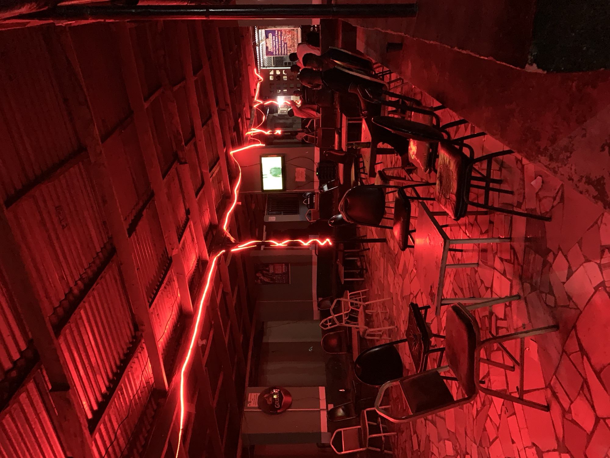 This image depicts a low-lit, open-air bar/restaurant in or near New Stadium, Nigeria. The space is illuminated by red string lights hanging from the corrugated metal roof and a red light source bathing the scene. A group of people, possibly customers, are seated on chairs at a long table towards the back of the space. A television screen is visible on the wall, with a poster or advertisement behind it. Various chairs and tables are scattered throughout the space, suggesting the bar's seating arrangement. The floor is tiled with irregular, broken pieces. A "Gold" branded sign is visible on a wall further back in the space.