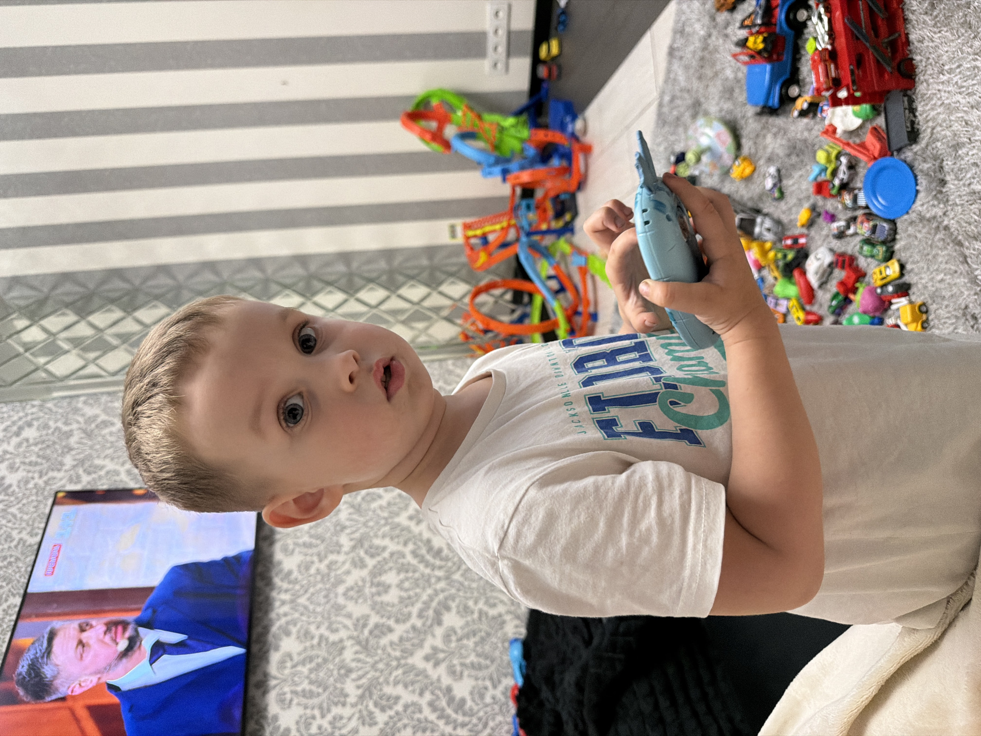 A young boy, with short brown hair and blue eyes, is holding a blue toy in his hands. He is wearing a white t-shirt with blue and green lettering. He is standing in a room with a grey carpet. In the background, there is a television screen displaying a man in a blue suit and tie, a toy race track, and various other toys. There is a striped wallpaper on the wall behind the boy. The room appears to be a living room or playroom. The image was taken indoors. The city of Bila Tserkva, Ukraine is not visible in the image.