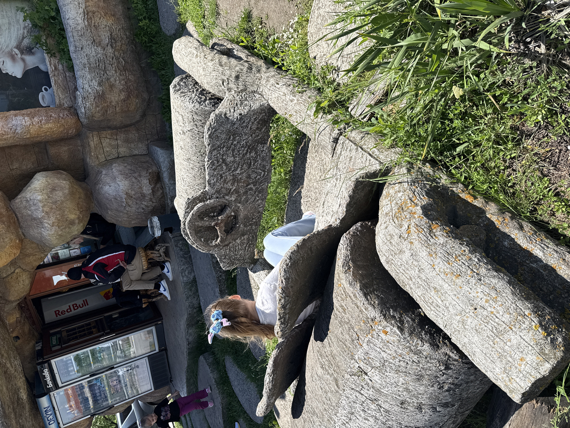 A ground-level shot, taken in Omurtag, Bulgaria, depicts a young girl in light blue jeans and a white t-shirt, partly obscured by a stone structure. The structure is composed of stacked, rounded stones resembling logs. The girl wears a blue and pink hair accessory. Beyond the stone structure, a grassy area is visible. To the left, a stone facade encloses a vending area displaying refrigerators filled with drinks. A "Red Bull" sign is prominently displayed above the refrigerators. People are near the vending area, appearing to be making purchases. The scene is bathed in natural sunlight, suggesting a clear day.