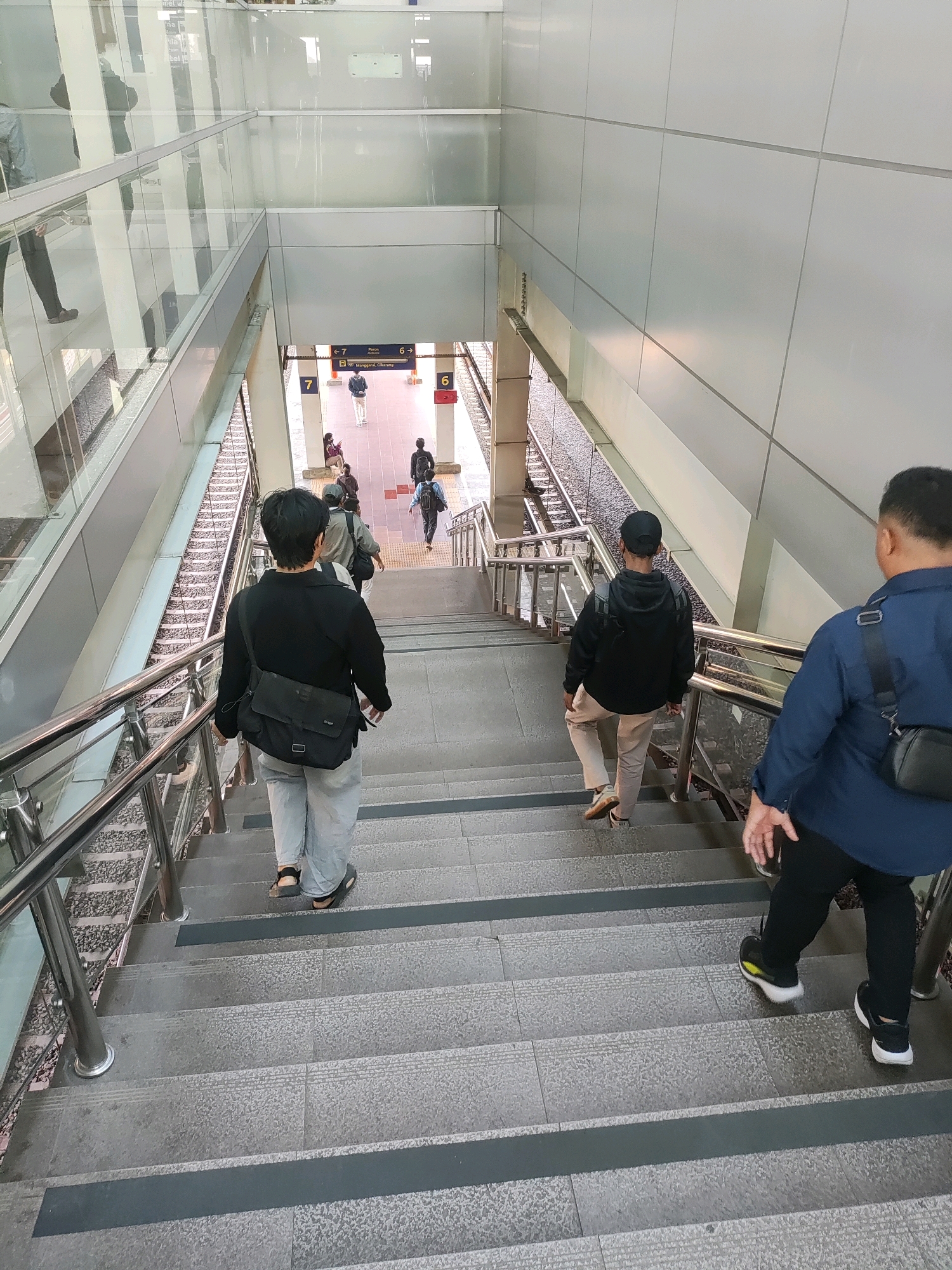 The image shows a downward view of a modern, multi-level train station in Bekasi, Indonesia, with people using the stairs. Several individuals are visible on the stairs and on a lower platform. One person in the foreground, viewed from behind, wears a black jacket, light grey pants, and a black crossbody bag, and is descending the stairs. Another person in a dark blue shirt and black pants, also viewed from behind, is descending the stairs ahead of the first individual. A third person, wearing a black hoodie and light-colored pants, is also going down the stairs further ahead. On the platform below, several other individuals are present, some seated and others standing. The station features contemporary architecture with glass railings, stainless steel handrails, and light-colored wall panels. Train tracks are visible to the right of the stairs, and signage indicating platform numbers and directions is present on the lower level. The scene appears to be during daylight hours.